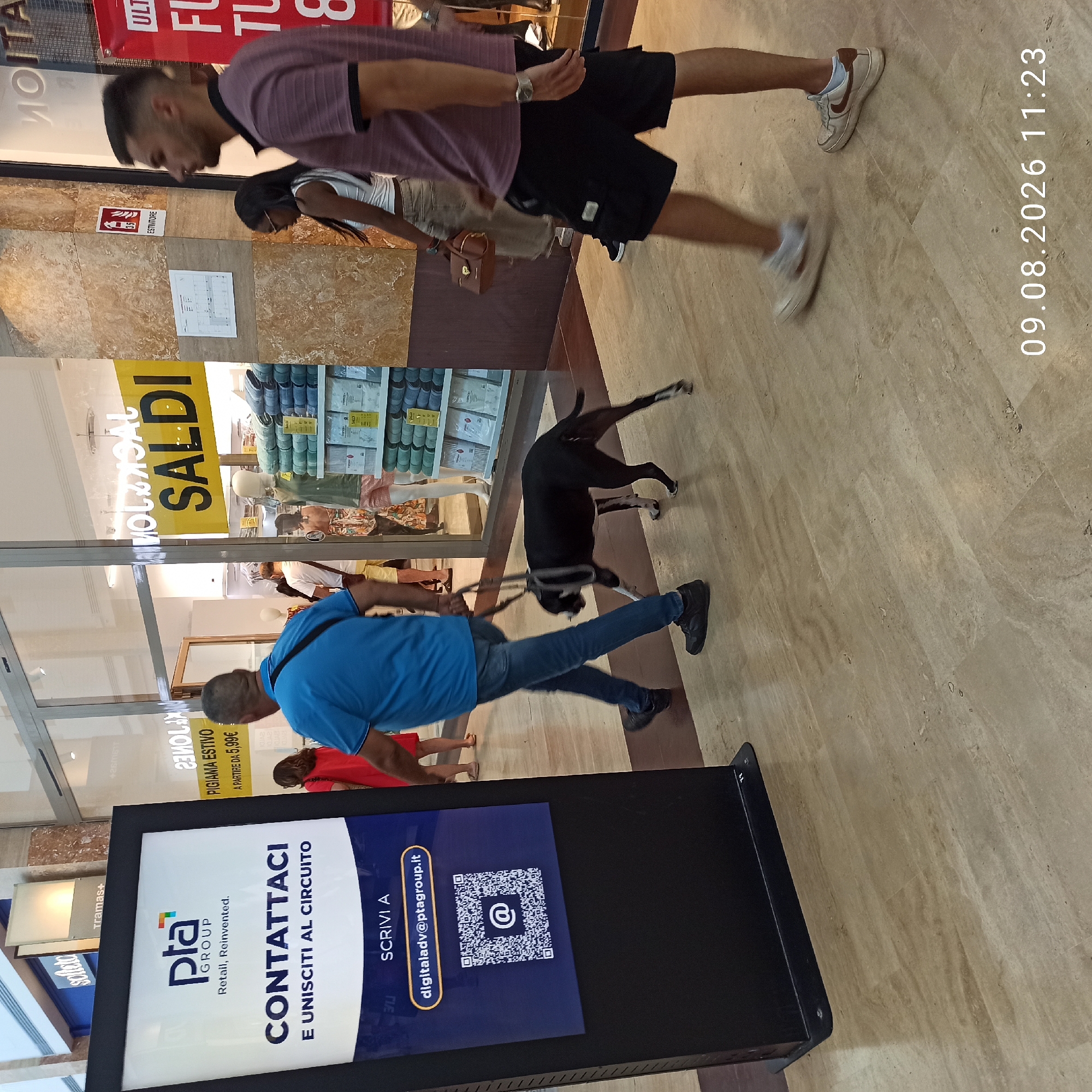 In Molfetta, Italy, a black and tan dog on a leash is being walked by a person wearing a blue t-shirt and jeans. The dog is walking in front of the person. In the background, there is a large digital display with the text "PTA GROUP" and "CONTATTACI E UNISCITI AL CIRCUITO" with an email address and QR code. To the right, several people are walking and browsing in what appears to be a commercial setting, with store windows displaying merchandise. A yellow sign with "SALDI" (sales) is visible in one of the windows. The floor is a light-colored marble tile. The timestamp "09.08.2026 11:23" is superimposed on the bottom right of the image.