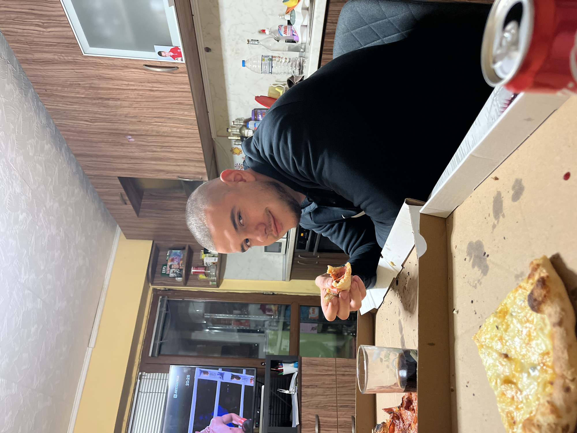 A man with a shaved head is holding a piece of pizza in his right hand while sitting at a table with a pizza box. The box contains pizza slices and an empty glass. A can of soda is partially visible in the upper right corner. In the background, a television displays a wrestling match. Through a window, other buildings are visible. Based on the presence of Bulgarian text on the television screen, it is likely that the location is in Bulgaria. Assuming the city to be Burgas is not verifiable from the visual data.