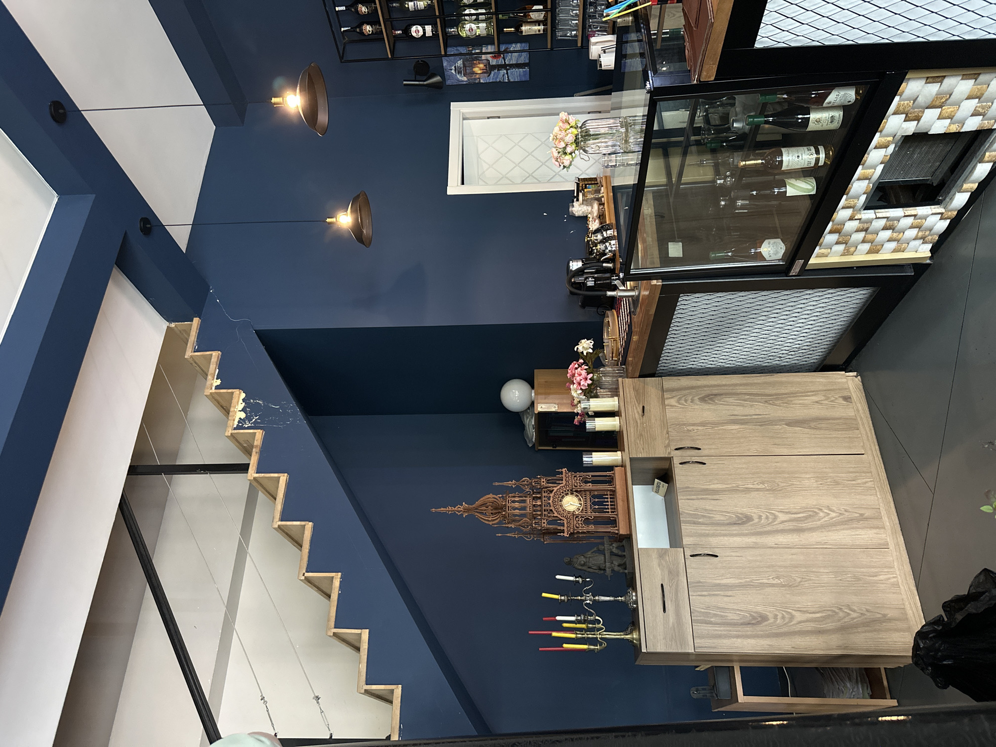 The image presents an interior view of what appears to be a business establishment, possibly a wine bar or boutique, situated in Marneuli, Georgia.
The decor features a modern aesthetic with dark blue walls contrasted by white architectural elements, most notably a staircase along the left side of the frame. Two pendant lights hang from the ceiling, illuminating the space.
On the right, a wooden cabinet with multiple drawers and cabinets stands against the wall. Above it, a display case with glass windows showcases wine bottles and glasses. Additional decorative items, such as flowers, candles, and an ornamental clock, are arranged on shelves and surfaces.
A visible rack of bottles mounted on the wall, reinforces the impression of a business serving alcohol, potentially wine. Overall, the scene depicts a well-maintained and thoughtfully decorated interior space in Marneuli, Georgia.