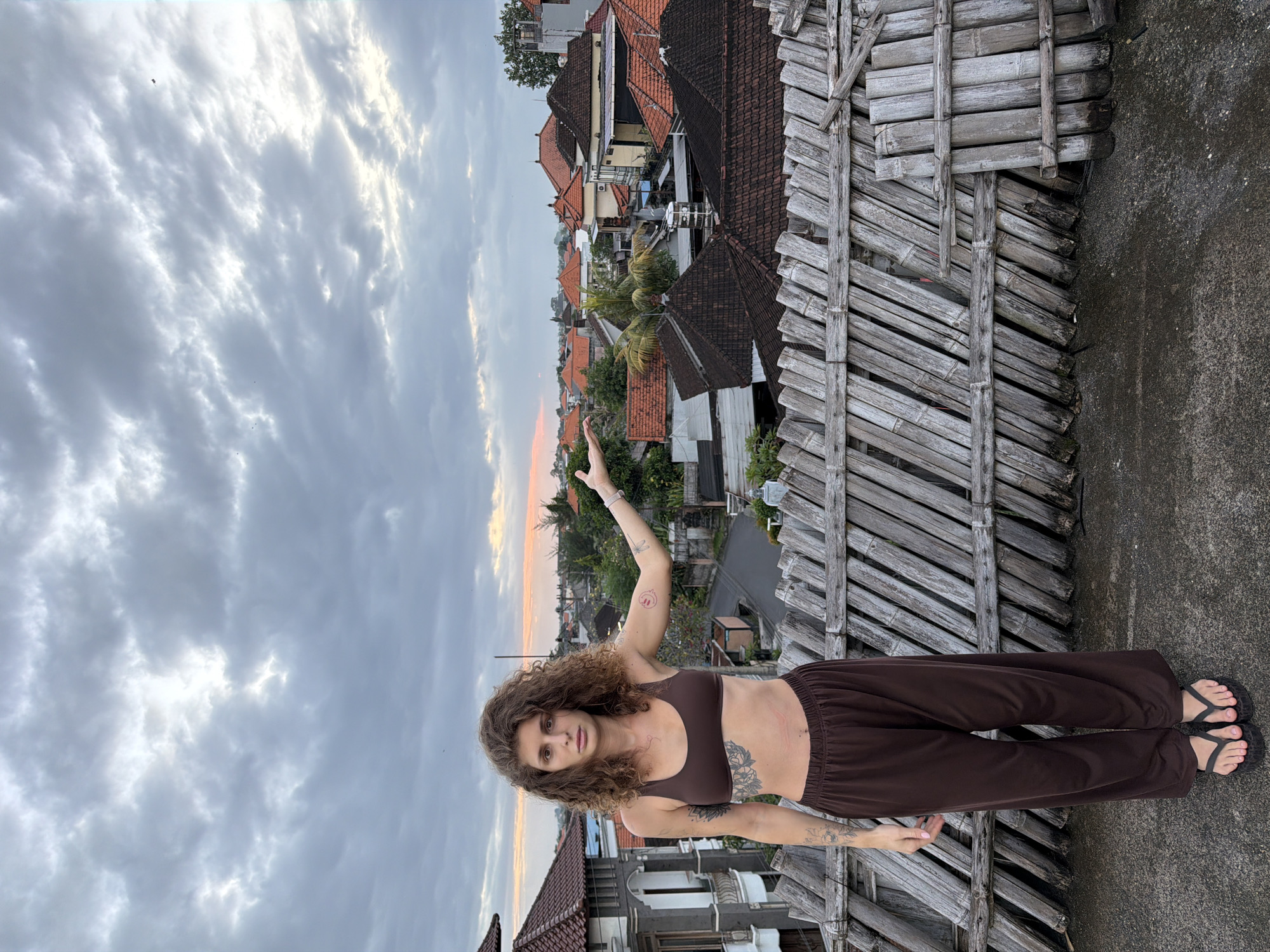 A young, slender person with curly brown hair stands on a rooftop. They are wearing a brown bra top and brown pants, and dark flip-flops. Tattoos are visible on their arms and torso. One arm is raised, pointing towards the background of the image. The rooftop is constructed of weathered wooden slats. Behind the person and the rooftop, there is a view of a city with closely packed buildings, mainly with orange tile roofs. A road is visible between some of the buildings. The sky above is cloudy, with the setting sun visible in the distance. The scene appears to be in North Kuta, Indonesia, based on the architecture and vegetation.