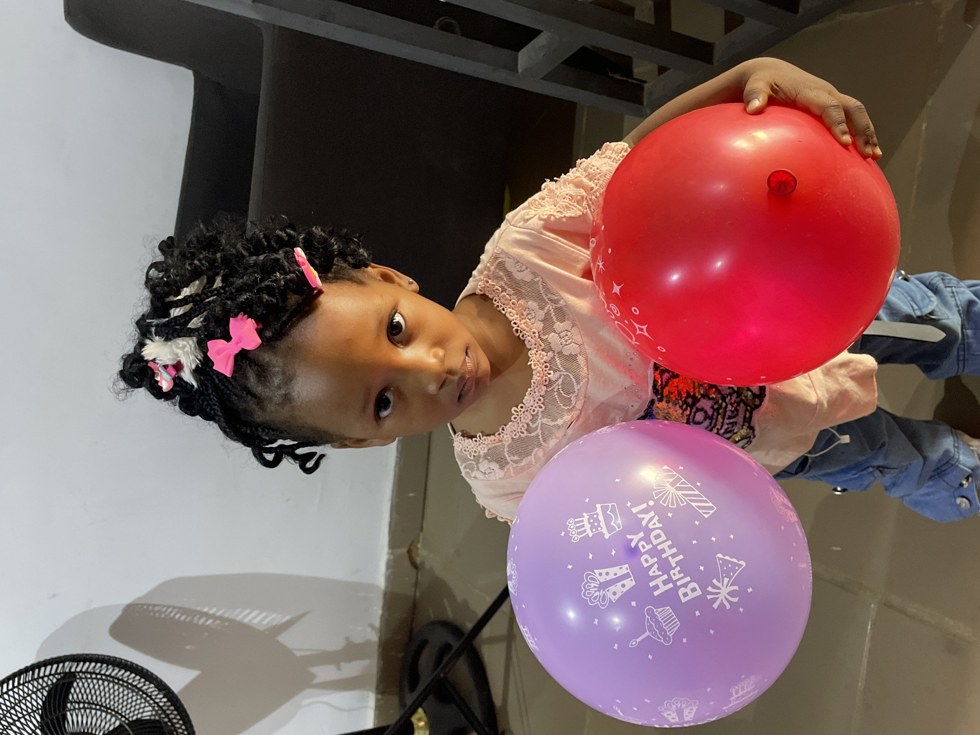 A young girl with braided hair, adorned with pink and white hair accessories, is holding two balloons. The balloons are red and purple, with the latter displaying "Happy Birthday!" in white lettering and cake designs. She wears a light pink, lace-trimmed shirt and blue denim bottoms. The scene includes a black oscillating fan in the lower left corner, a dark object above, and a partially visible interior setting. The location is Barakin Akawo, Nigeria.