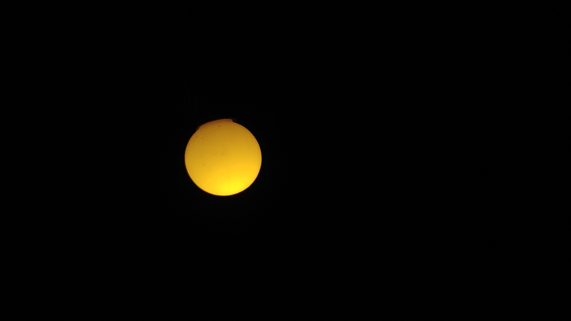 A bright yellow, spherical light source is centrally located against a completely black background. The light source appears to be emitting a strong glow, with subtle variations in its color and texture, suggesting it could be a lamp or a celestial body viewed through a camera lens. The scene is devoid of any other objects, people, or environmental details.