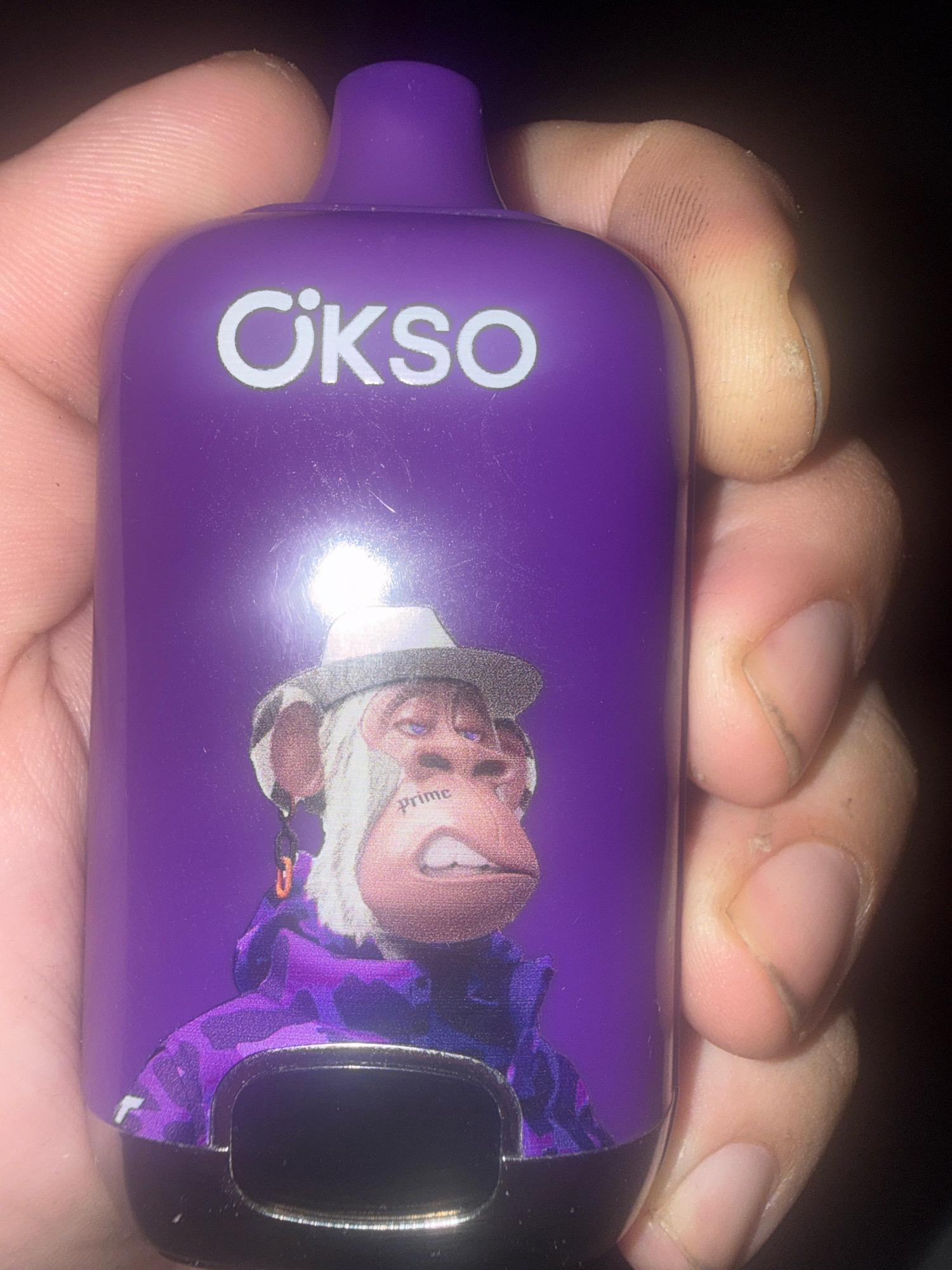 A close-up shot shows a person's hand holding a purple vaping device. The device has the brand name "OKSO" printed in white, and a graphic of a stylized ape wearing a hat and a purple camouflage jacket. The ape graphic also features the word "prime" tattooed on its cheek. The background is dark and out of focus. There are no discernible features that place this image in Gorzow, Poland.