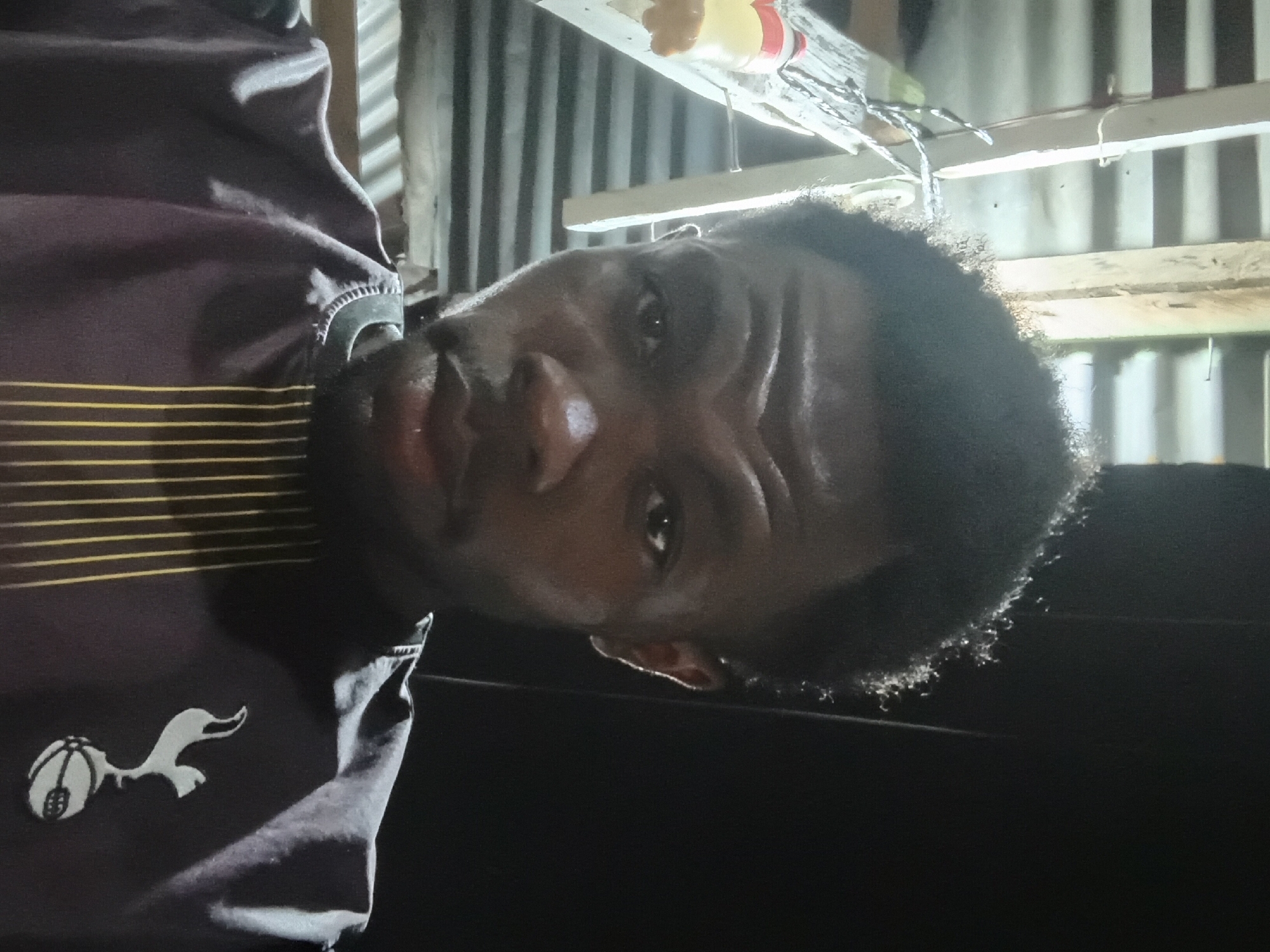 A close-up, waist-up, front-facing portrait of a dark-skinned male is captured in a dimly lit indoor setting. The man has short, dark, curly hair that appears to be styled upwards and slightly to the right. He has prominent eyebrows, dark eyes, a wide nose, and full lips. His expression is neutral, with his gaze directed towards the camera.
He is wearing a dark purple t-shirt with a V-neck collar adorned with white stitching. Across the front of the shirt, from the collar down to the chest, are vertical yellow lines of varying thickness. On the left side of the chest, a white emblem of a rooster on a ball is visible, suggesting it is a sports team's logo.
The background is dark and appears to be a rustic, possibly unfinished interior. To the left of the man, a light-colored wooden structure is visible, possibly part of a shelf or support beam, with some indeterminate items hanging from it. A plastic bottle with a red cap and yellow liquid is partially visible on this structure. The background also shows horizontal ribbed metal sheeting, typical of corrugated iron, which may form a wall or roof.
The lighting in the image is uneven, with strong highlights on the man's forehead, nose, and cheekbones, indicating a light source from the front and slightly above. The overall atmosphere is informal and personal. The location is identified as Jalingo, Nigeria, suggesting this image was taken within that city.