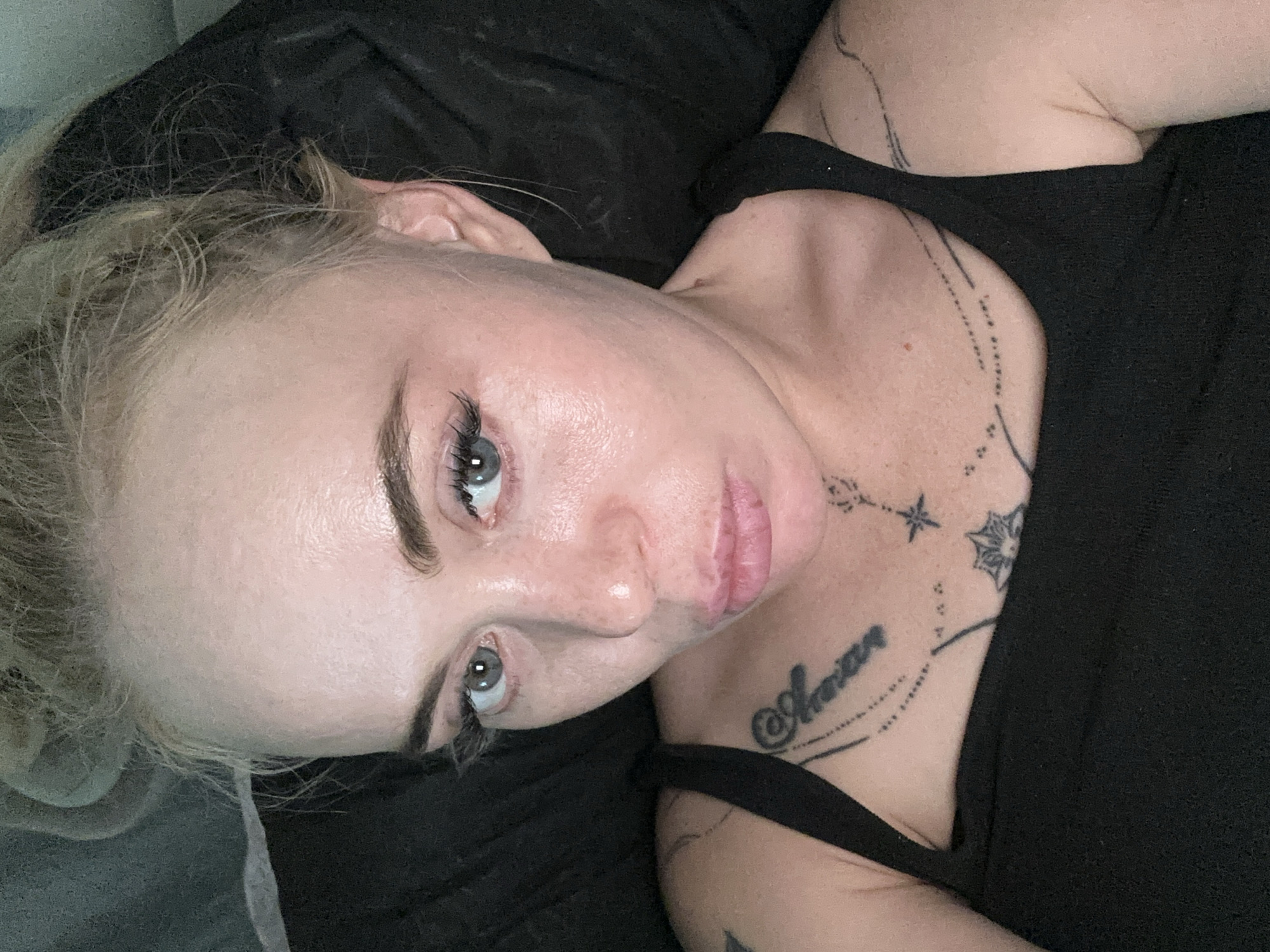 A woman with blonde hair pulled back and wearing a black tank top is shown from the chest up. She has blue eyes, prominent eyelashes, and visible freckles. There are tattoos on her chest and shoulder. The background is dark and appears to be a fabric surface. The image is a close-up, self-taken photograph.