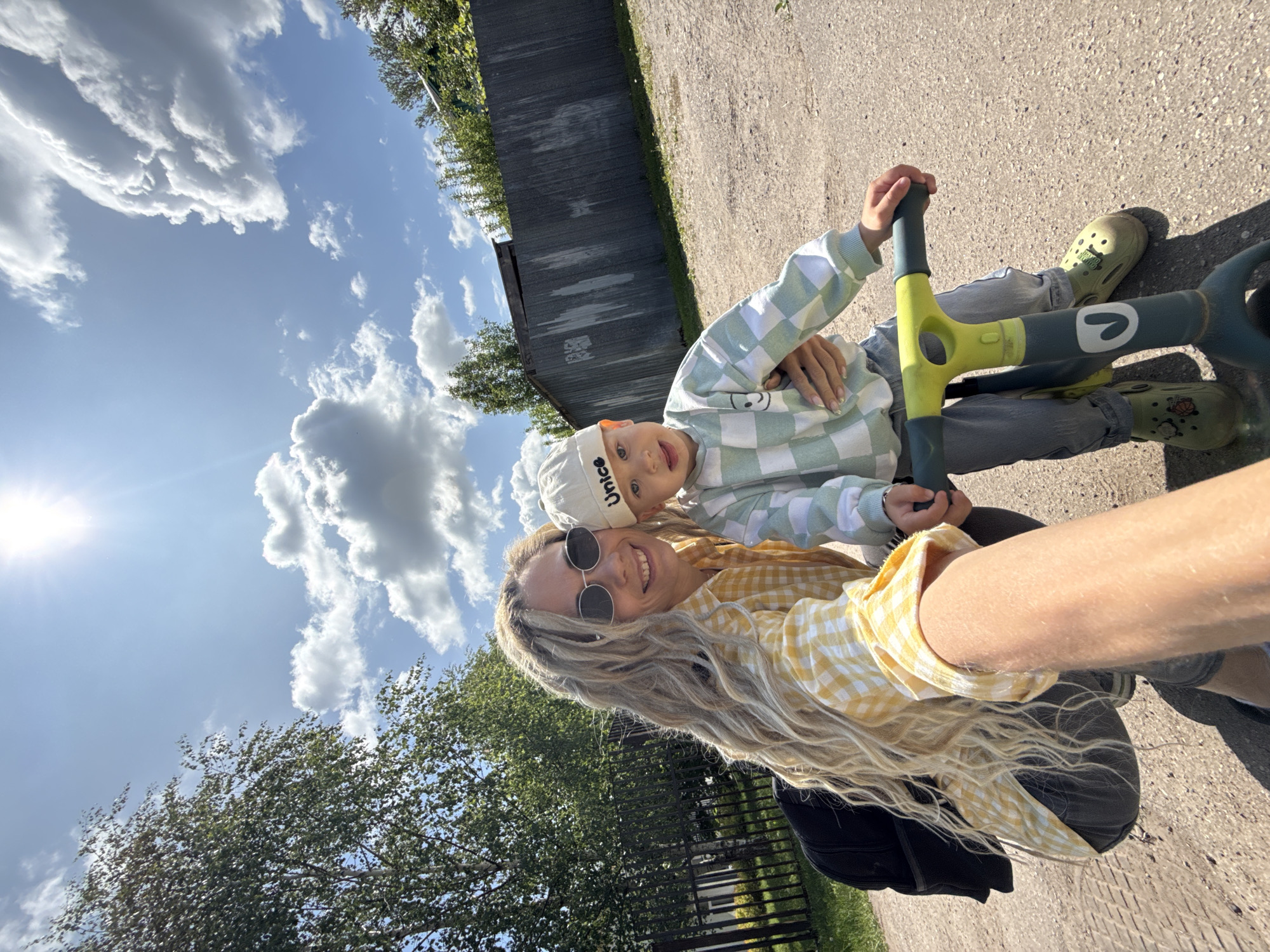 The image captures a diagonal selfie-style shot of a woman and a young child outdoors on a sunny day in Rusanovo, Russia. The camera perspective is tilted, making the paved ground appear on the right side of the frame and the sky on the left.
In the foreground, a woman with long, wavy blonde hair and black sunglasses smiles towards the camera. She wears a yellow and white checkered long-sleeved shirt. Her left arm, holding the camera, extends into the lower right corner of the frame.
Beside and slightly behind her is a young child, likely a boy, looking at the camera with a slight smile. He wears a white beanie with the word "NICE" visible on the front, a light green and white checkered sweatshirt featuring a small smiley face graphic, and light blue jeans. His feet are clad in olive-green Crocs-style sandals adorned with small charms. The child is seated on a gray and lime green balance bike or scooter, which features a white logo on its frame.
The setting appears to be an outdoor paved area, possibly a path or driveway, visible on the right side of the image. Behind the subjects, a dark gray wooden or metal fence stretches across the mid-ground. Lush green trees are visible on the left and right edges of the frame, and also in the distance behind the fence. The sky, filled with bright sunshine and scattered fluffy white clouds, dominates the upper left portion of the image, indicating clear and pleasant weather. Both subjects' smiles suggest happiness and enjoyment of their outdoor activity.