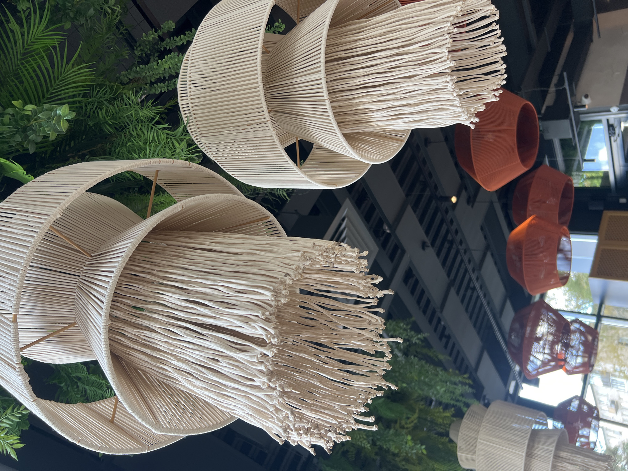 This is a low-angle, eye-level shot of several hanging pendant lights and artificial greenery, likely indoors in a commercial establishment in Veliky Novgorod, Russia. In the foreground, two large, tiered lampshades made of cream-colored macrame or woven cord hang prominently. The shades have a conical shape with multiple layers of the cord creating a fringed effect at the bottom. Behind these, other similar cream-colored lights, also tiered, are visible to the left. To the right and in the background, several bell-shaped lampshades in a vibrant orange or reddish-orange color hang. Artificial green foliage, including ferns and succulents, is visible in the upper portion of the image, suggesting a decorative or thematic design. The ceiling appears to be a dark, industrial style with exposed beams and lighting fixtures. Large windows are visible in the background, showing a blurred view of buildings and possibly daylight outside. There are no people visible in the image.