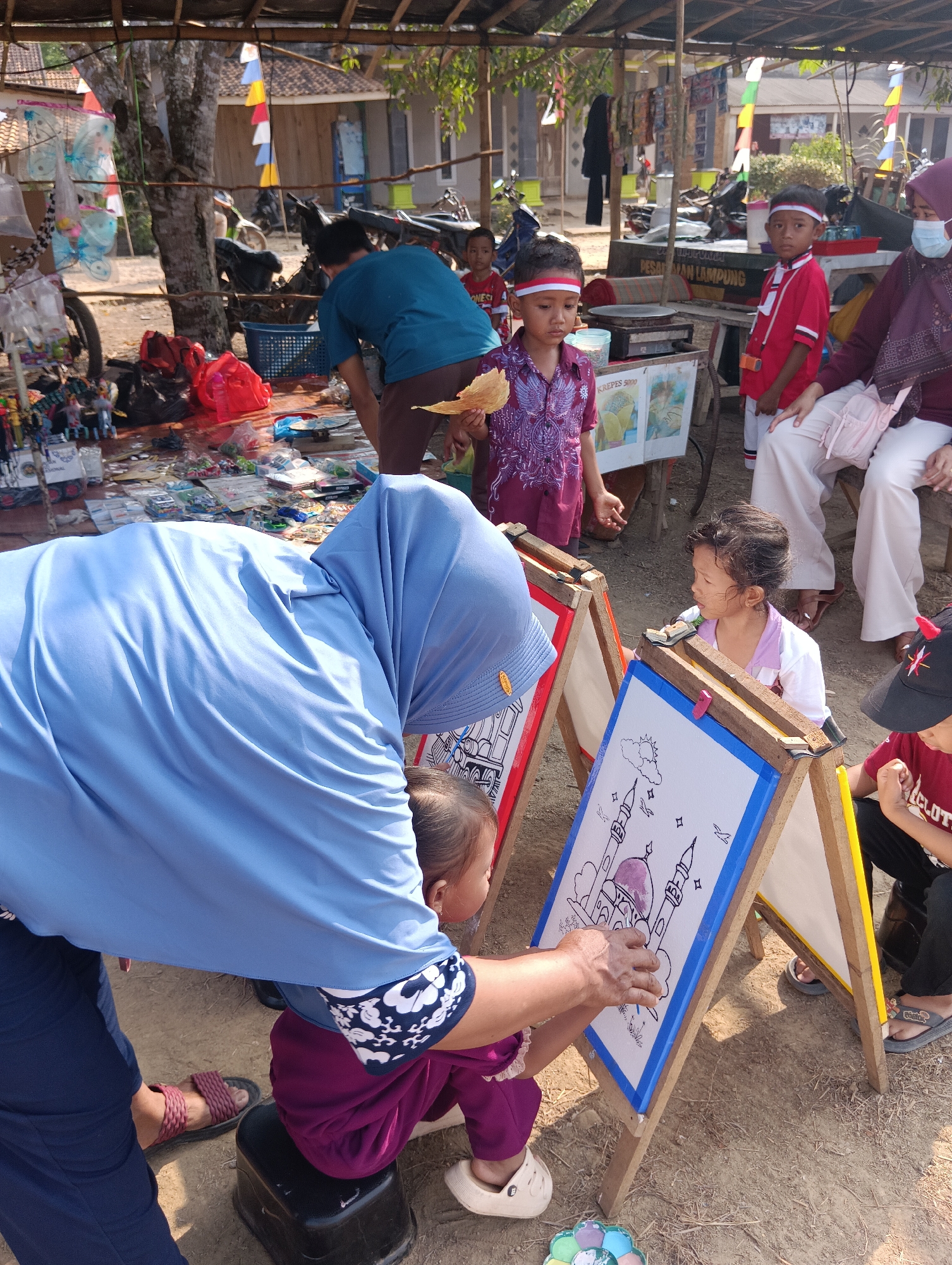 The image depicts an outdoor scene in Pematang Jaya, Indonesia, likely a market or community gathering. In the foreground, a woman wearing a blue hijab and a matching blue garment is bending over to assist a young child who is sitting on a black stool. The child is wearing a purple outfit with white trim and is focused on drawing on a white board placed on a wooden easel. The drawing on the board appears to be a mosque with minarets. Another child, also drawing, is visible to the right, wearing a white and purple shirt. Behind them, several other people are present, including children wearing red and white headbands, suggesting a festive occasion. A woman in a maroon headscarf and mask is seated, and other individuals are standing. The environment is dusty with dry grass and scattered items. In the background, there are stalls with various goods, motorcycles, and buildings, hinting at a bustling marketplace. Flags are visible, adding to the sense of a community event. The overall atmosphere appears to be lively and engaged.