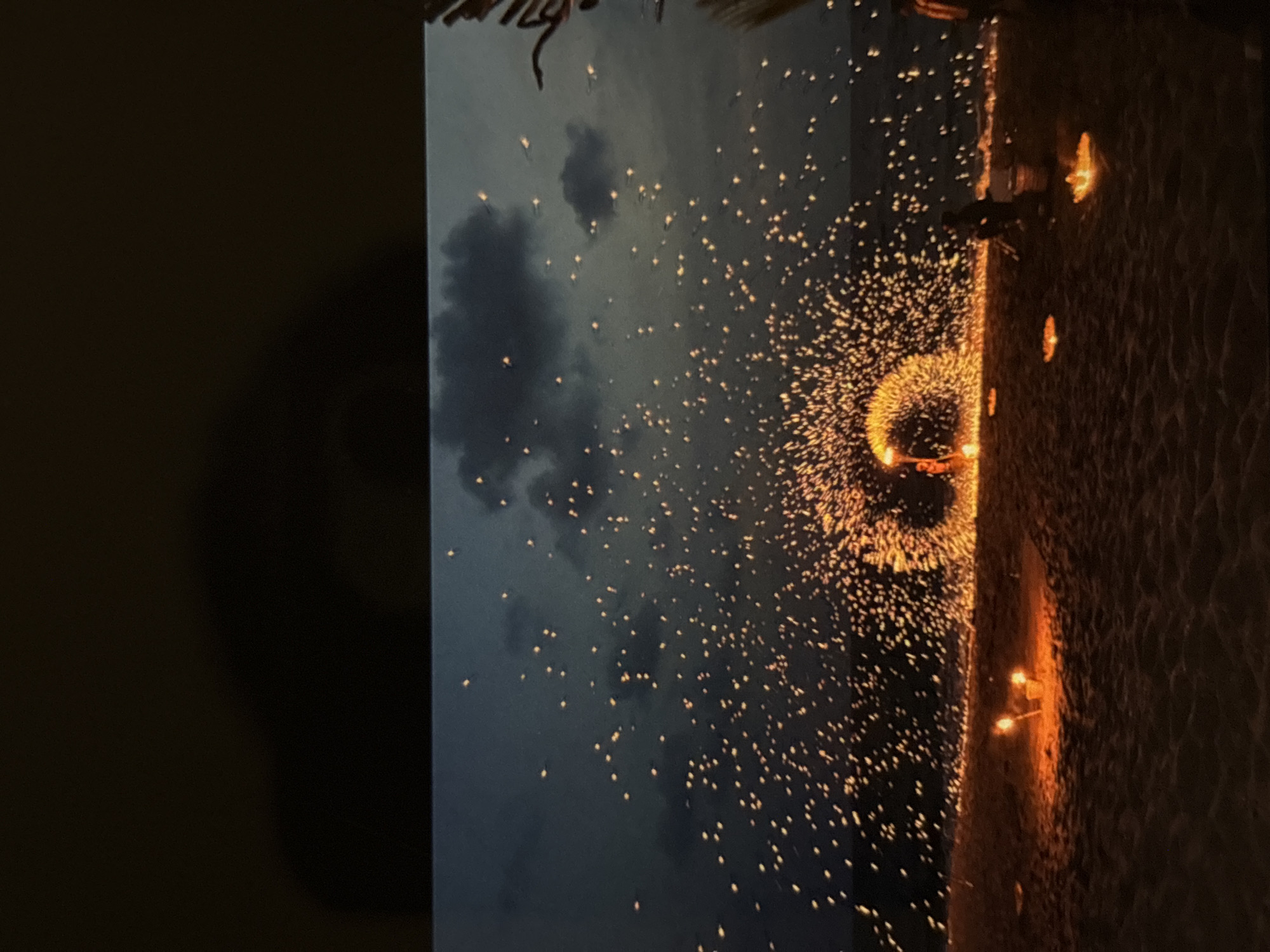 The image depicts a fire performance on a beach at dusk, with the ocean visible in the background under a dark, cloudy sky. The sand is illuminated by numerous glowing embers scattered across the scene, creating a magical effect. In the center of the image, a performer is creating a large ring of fire, with sparks flying outwards. There are also several smaller fires or lanterns scattered on the sand in the foreground and midground. Two figures are partially visible in the distance, one standing near the fire performer and another to the far right. The overall atmosphere is one of spectacle and entertainment, likely taking place in a tourist area such as Chalong, Thailand, known for its beachside activities.