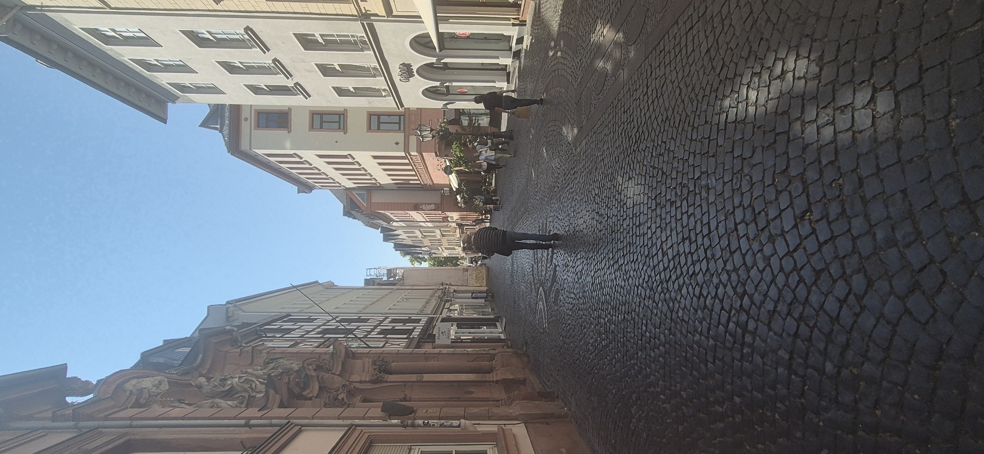 This is a vertical, eye-level, full shot of a cobblestone street in Mainz, Germany. The street is lined with historic European buildings, some with ornate facades and others with more simple, multi-story designs. The sky is clear and blue. Several people are walking in both directions on the street. In the foreground, a person wearing a striped shirt and blue jeans walks away from the camera. Further down the street, another person with dark hair walks toward the camera carrying a bag. Other individuals are visible in the distance, some appearing to be shoppers or patrons of the shops lining the street. Some of the storefronts have awnings. The cobblestone pavement is textured and appears worn, with shadows from the buildings cast across it. The buildings on the left have classical architectural details, while those on the right are more modern in style. The overall impression is of a lively, European city street on a sunny day.