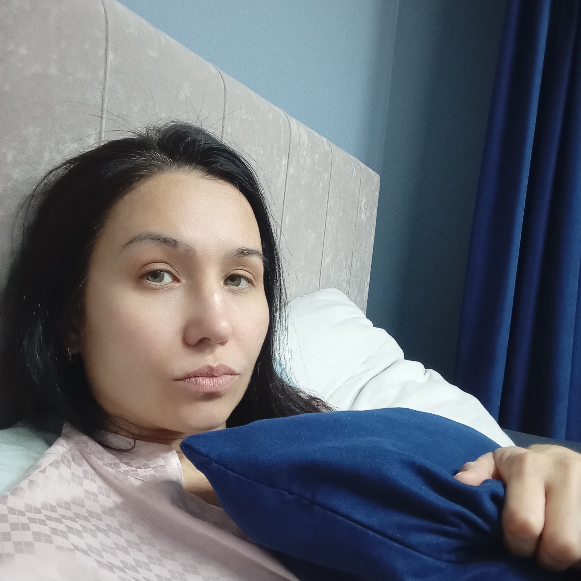 A woman with black hair and light eyes is lying in bed, holding a blue pillow. She is wearing a pink and white patterned top. The headboard is light gray. Behind her is a blue wall and dark blue curtains. The location in the city of Pervomaysky, Russia, is not discernible from the image.
