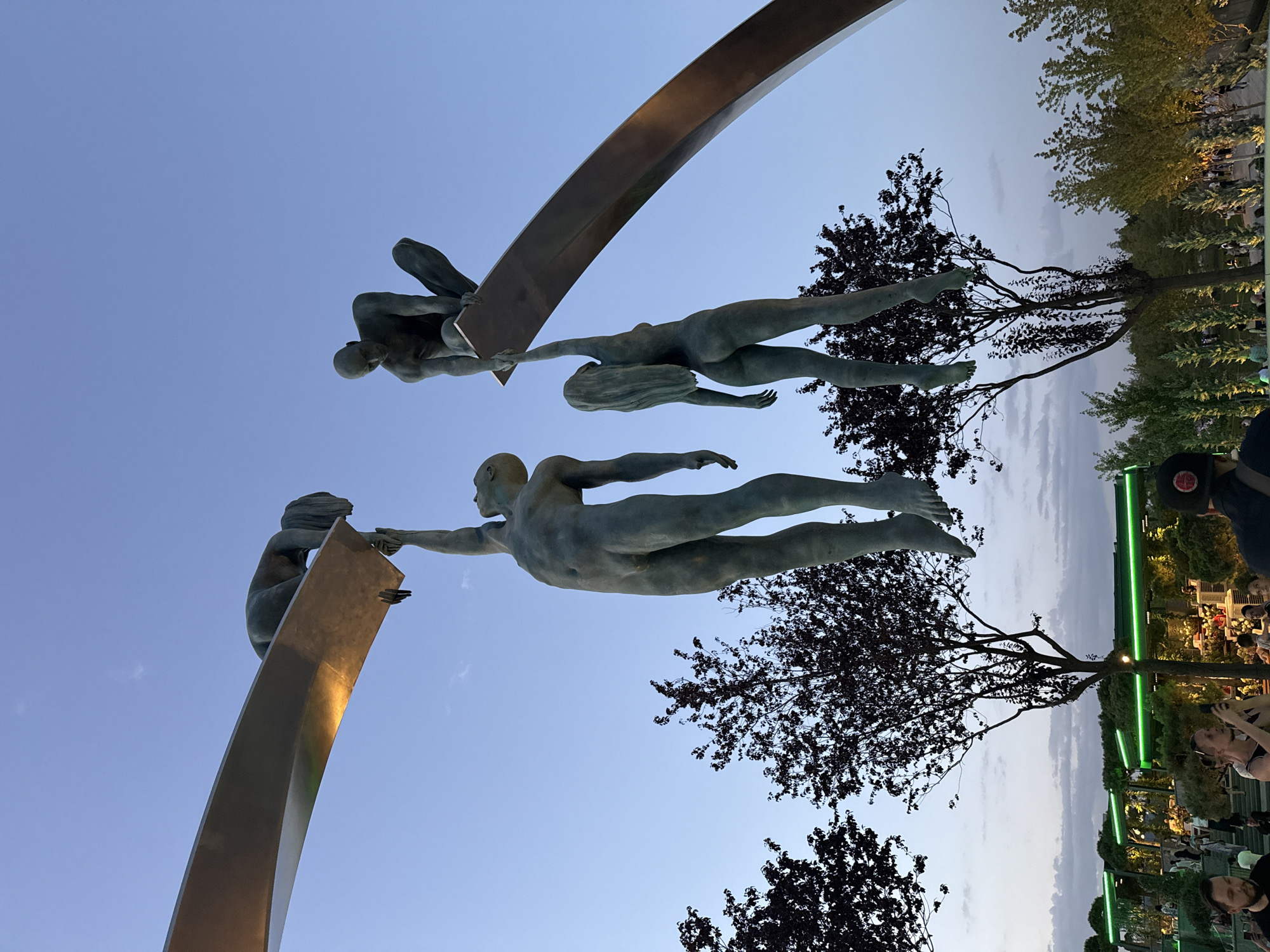 The image shows a bronze sculpture of four human figures in a park-like setting in Krasnodar, Russia. Two figures are suspended in mid-air, reaching towards each other. One figure is crouched on a curved metallic beam, and another figure is reaching up to it from below. Below the sculpture, there are trees with dark foliage, and in the background, there are illuminated structures resembling cafes or restaurants with green lighting. Several people are visible in the lower part of the image, some of whom are looking at their phones. The sky is a clear blue, indicating it is likely daytime or early evening.
