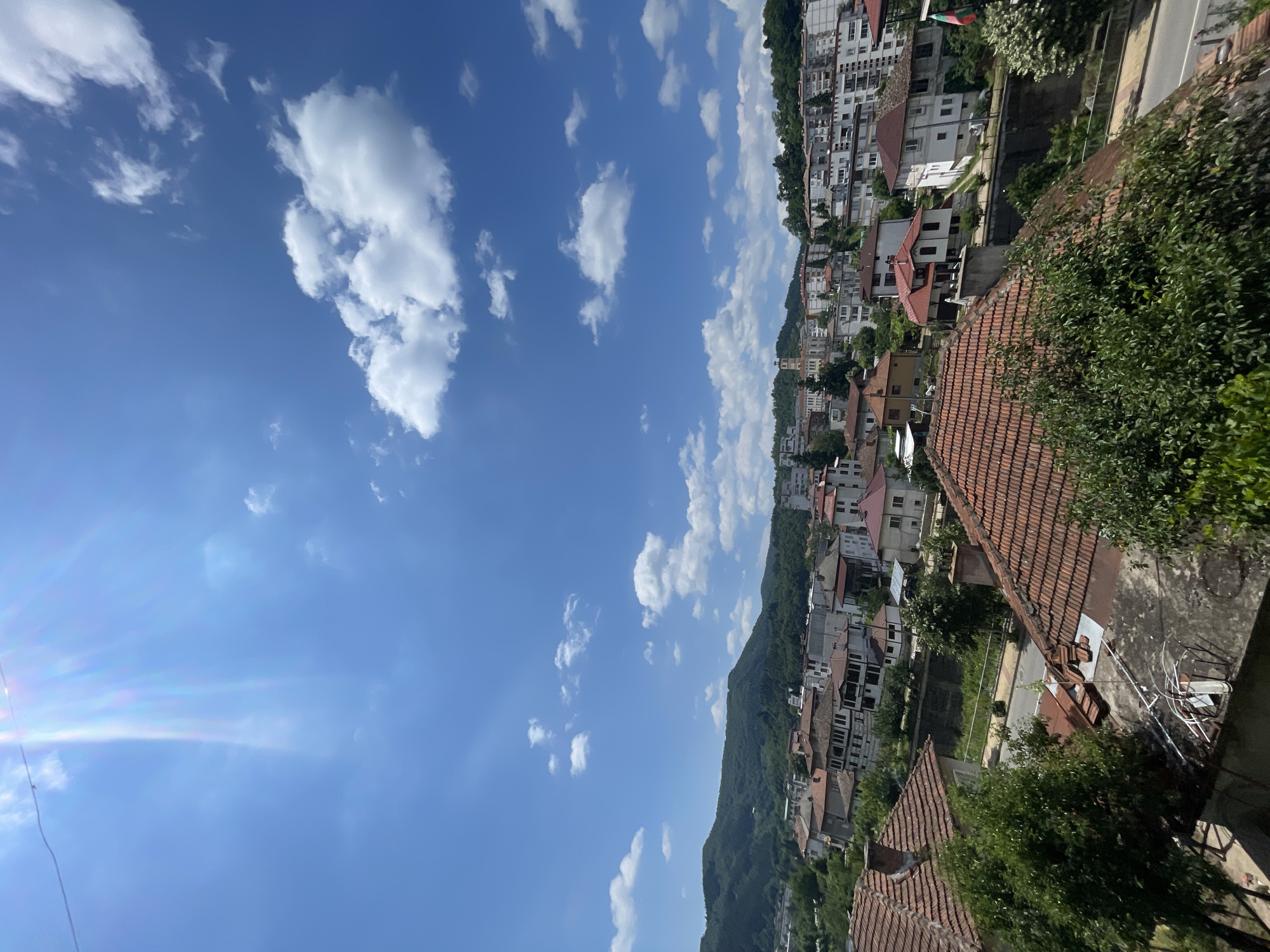 This media file presents a scenic view of a town nestled on a hillside under a bright, partly cloudy blue sky. The architecture is a mix of traditional tiled roofs and more modern, multi-story buildings, creating a sense of depth and history. Green trees and foliage are interspersed throughout the town, cascading down the slopes of the hill. The sun is shining, indicated by the bright illumination of the buildings and the presence of lens flare in the upper left portion of the image. The clouds are scattered and white, suggesting fair weather. The perspective is from a high vantage point, looking down and across the town, offering a panoramic vista. There are no people or specific activities depicted, making it a still-life landscape. The overall impression is one of a peaceful and picturesque setting, likely during the daytime.