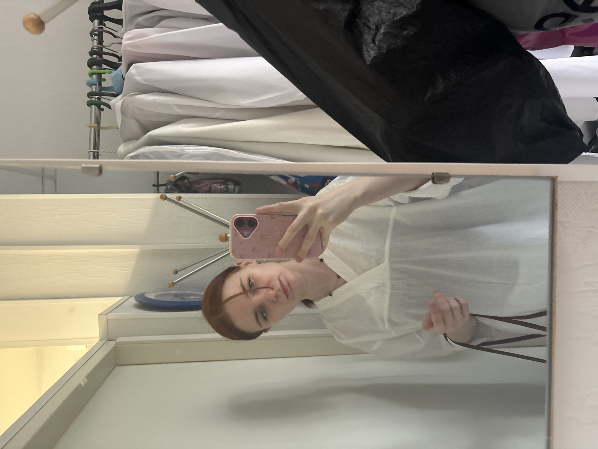 A mirror reflects a woman taking a selfie. She holds a pink phone with both hands. Her hair is reddish-brown, and she is wearing a white blouse. The background visible through the mirror shows a set of stairs, a blue plate, and what appears to be a white wall. Outside of the mirror's reflection is a clothes rack with hangers, stacked white fabric, and a black bag. The location is not identifiable from this image.