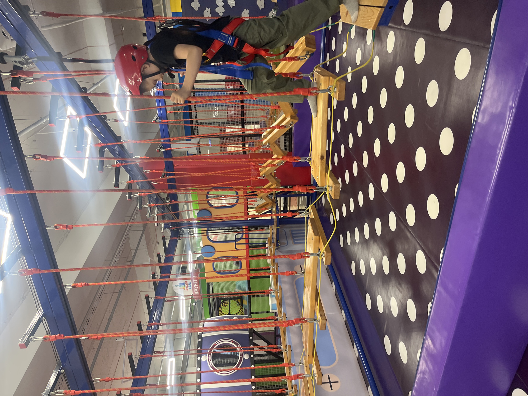 The image shows a person on a rope course in what appears to be an indoor entertainment facility. The person, wearing a red helmet, black shirt, and olive green pants, is attached to a safety harness and is navigating a series of wooden platforms and ropes suspended above the ground. The flooring underneath has a pattern of white circles on a dark background. The course includes other structures and colorful play areas. "Джунгли", visible on one of the play area structures, suggests that the photo may be located at Джунгли Kaspiysk in Russia.