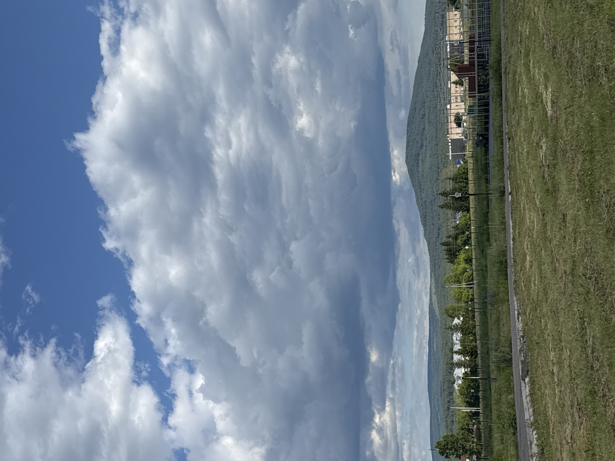 The image depicts a wide-angle view of a rural landscape under a cloudy sky. In the foreground, a mown grassy field extends towards a paved pathway. Beyond the pathway, a fence line runs horizontally across the scene, separating the field from a row of buildings and trees. The buildings appear to be residential or institutional, with several structures featuring light-colored facades and pitched roofs. Behind these buildings, a dense forest covers rolling hills, leading to a prominent, tree-covered mountain in the distance. The sky is filled with dramatic clouds, ranging from bright white to dark gray, suggesting an overcast or impending weather change. The overall impression is one of a serene, possibly remote, location.