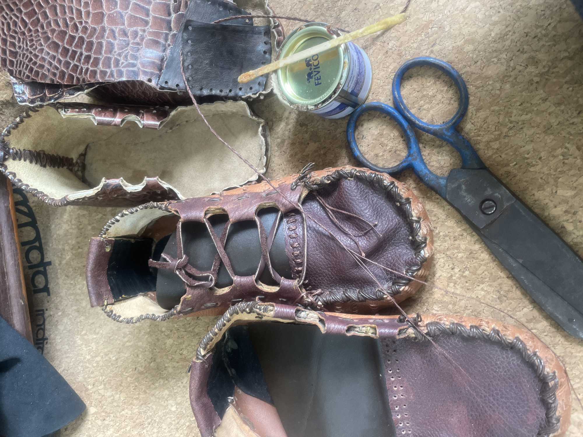 The image displays a close-up overhead view of a collection of handcrafted leather shoes and crafting supplies laid out on a cork-like surface. Two partially assembled leather shoes, a moccasin style with visible stitching and lacing, are prominent in the foreground. To the right, there are pieces of darker, textured leather, possibly intended for shoe parts, and a small tin of "FEVICO" adhesive. A wooden applicator stick coated in adhesive rests on the tin. A pair of blue-handled scissors and a spool of thin brown thread are also visible. In the background, a dark fabric item and a piece of wood with text are partially visible. The overall impression is of a workspace where leather goods are being handmade. The location in Bhagsu, India is not discernible from the image.