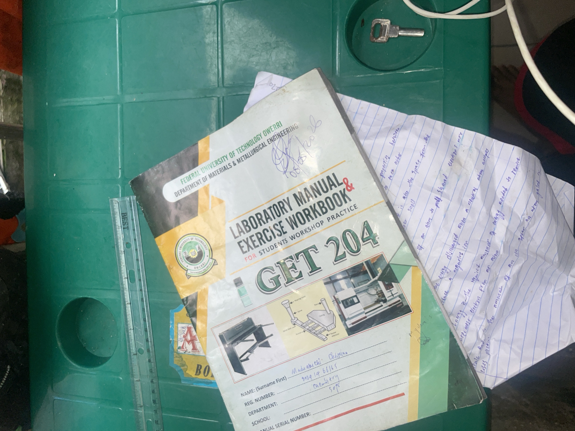 A laboratory manual for students titled "GET 204" and "LABORATORY MANUAL FOR STUDENTS WORKSHOP PRACTICE" is placed on a green surface, likely a table, at the Federal University of Technology, Owerri, Nigeria. The manual is open, revealing handwritten notes and diagrams. A ruler labeled "BEST" is positioned above the manual. The scene suggests an academic or study setting.