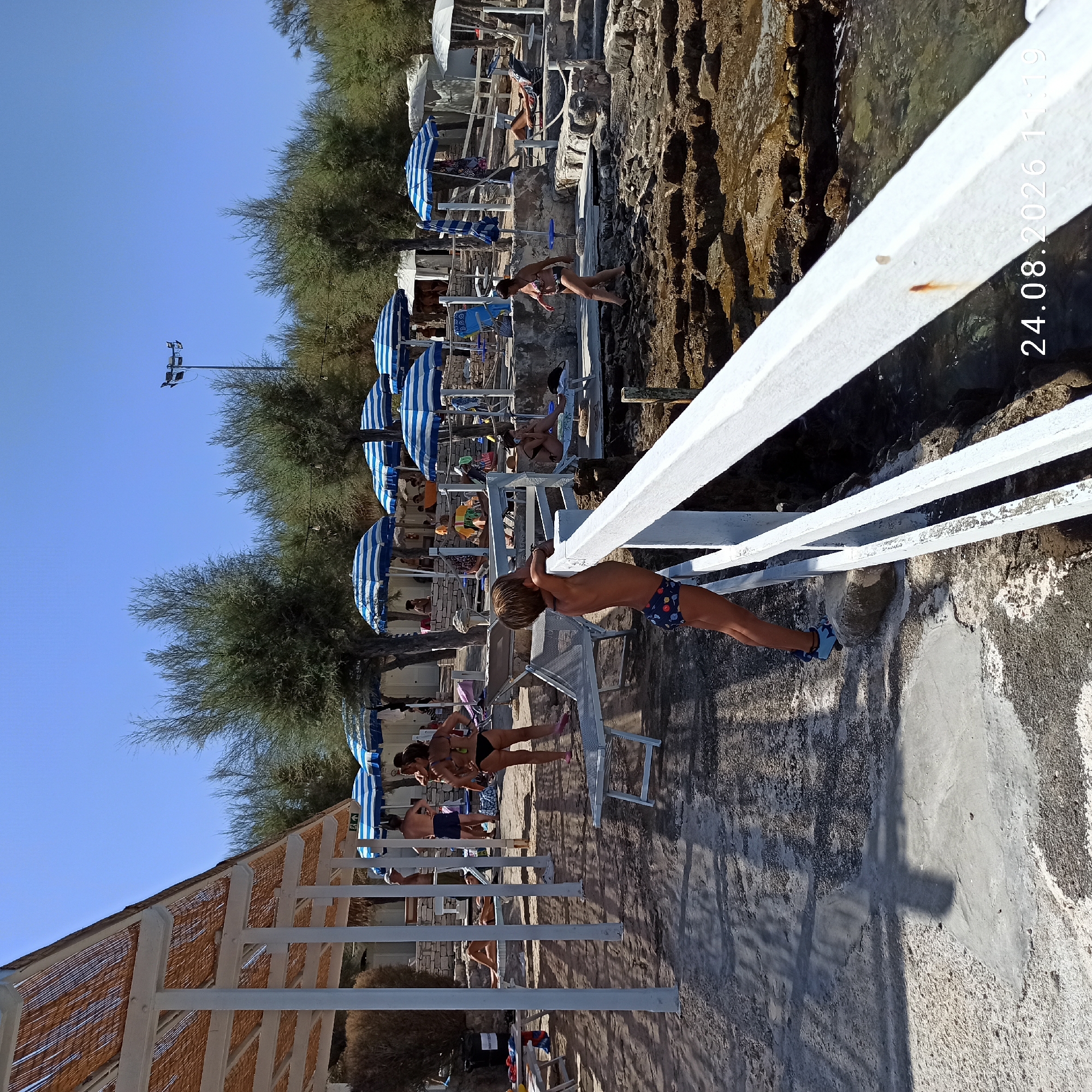 The image depicts a sunny day at a beach resort in Molfetta, Italy, likely the Lido Torre Cono. In the foreground, a young boy in patterned swim trunks leans on a white railing, looking out towards the sea. Behind him, scattered beachgoers are visible. Some are sunbathing on lounge chairs, while others are walking or standing near striped blue and white umbrellas. Structures resembling small cabanas line the background, partially obscured by trees. The ground is a mix of sand, concrete, and rocky shore. The sea is calm and clear, with visible rocks in the water. The timestamp "24.08.2026 11:19" is overlaid on the bottom right of the image.