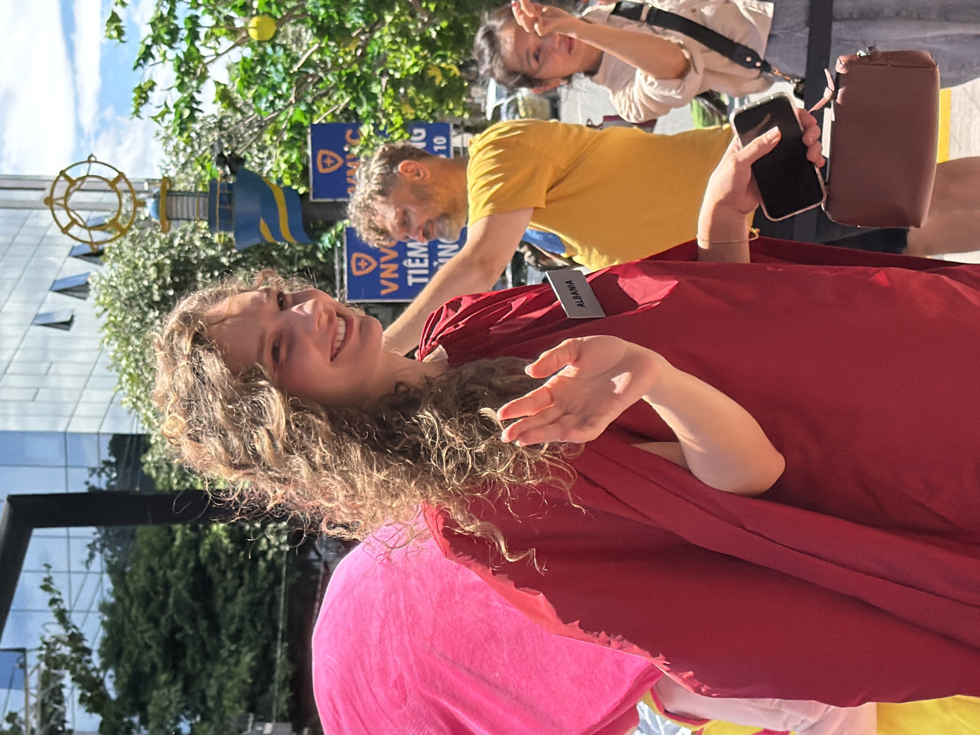 A young woman with curly blonde hair and a red dress stands in the foreground, smiling and waving to the camera. She is wearing a rectangular badge on her dress that reads "ALBANIA". To her left, a pink fabric is visible. Behind her, a man in a yellow t-shirt looks to his right. To the far right, a young girl with dark hair points to something off-camera. In the background, there is a blue building with glass windows, trees, and street signs. A decorative lamppost with a yellow circular emblem is also visible. The scene appears to be a public outdoor area, possibly a street or promenade in Nha Trang, Vietnam, given the context of the building and signage.