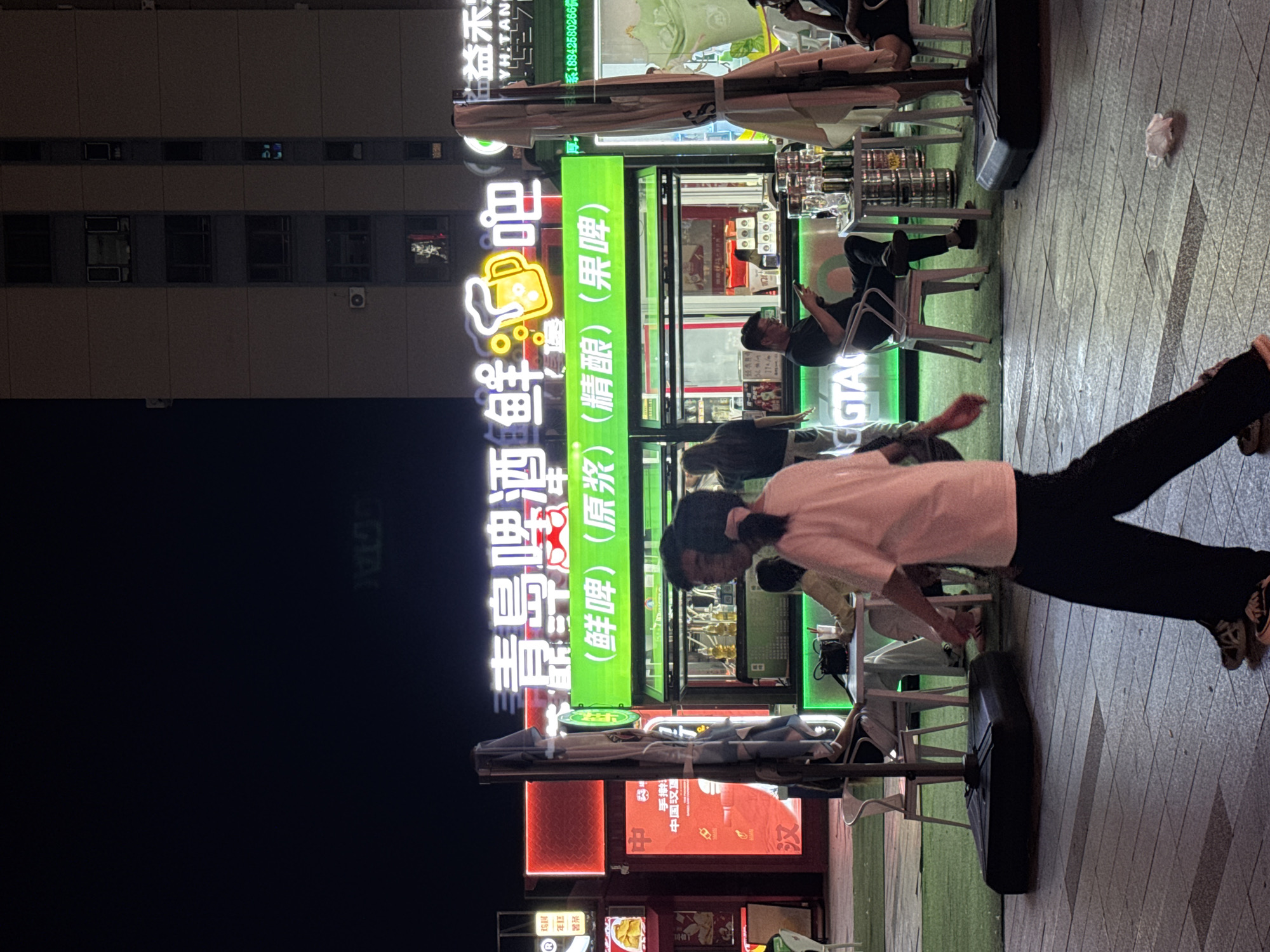 The image depicts a nighttime street scene in Shenyang, China, likely in an area with outdoor dining and entertainment. A brightly lit green stall with neon signs advertising "Qingdao Beer Bar" dominates the center, offering various types of beer. Several people are visible. In the foreground, a person walks away from the camera, wearing dark pants and a light-colored shirt. Further back, a man sits at a table, looking at his phone. Other patrons are seated at tables, some with drinks. Behind the stall, a tall building is visible with lit windows, suggesting it's an apartment or office building. The ground is paved with interlocking stones, and artificial turf is laid out in front of the stall. Umbrellas are set up to provide shade. The overall atmosphere is lively and suggests a popular spot for evening leisure.