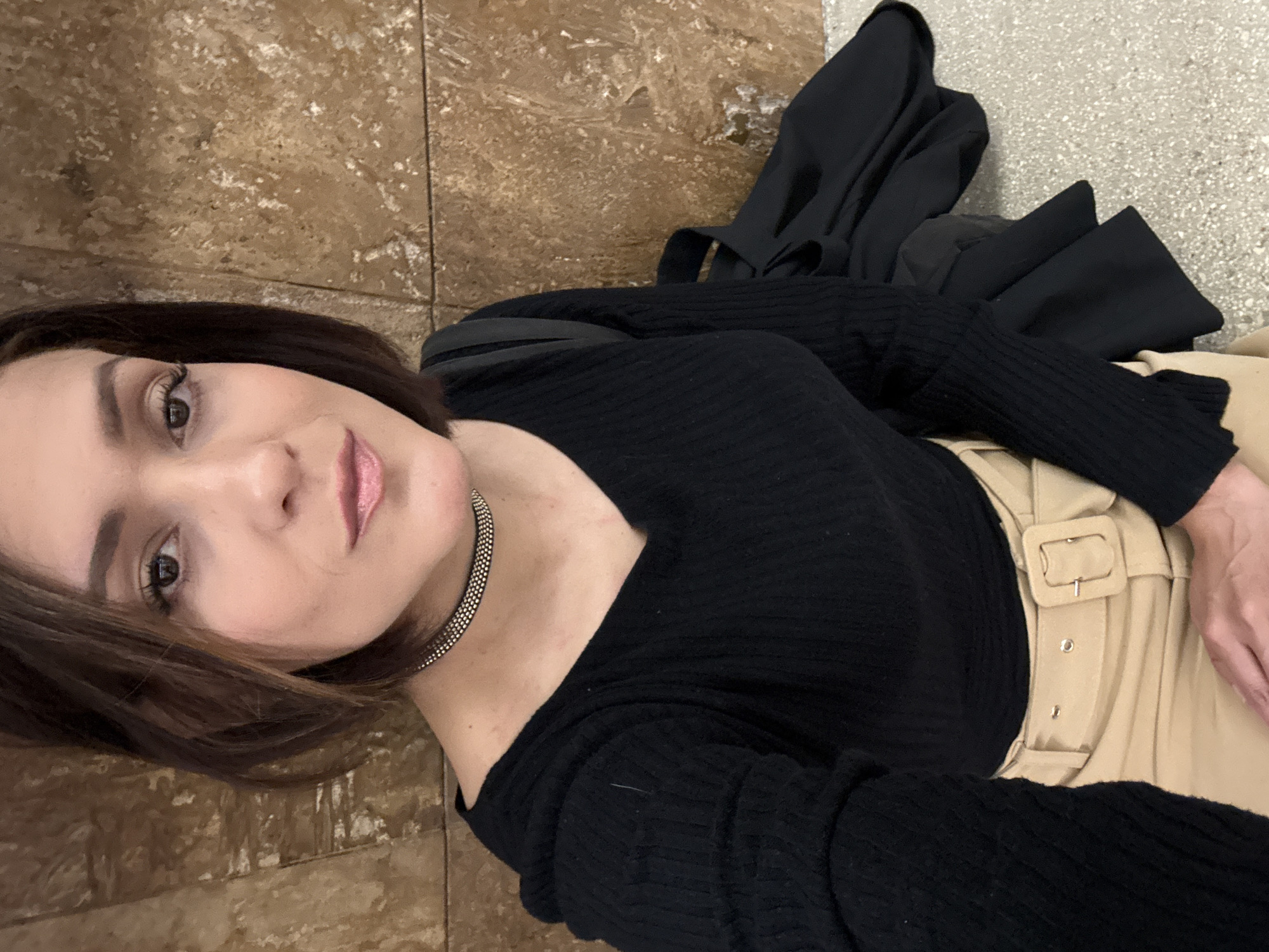 A woman with shoulder-length brown hair and fair skin is taking a selfie. She is wearing a black long-sleeved shirt and a choker necklace. She also wears a beige belt with a prominent buckle. A black jacket is draped behind her. The background consists of brown patterned tile flooring and a light grey surface. The location is unknown and cannot be determined from the image.
