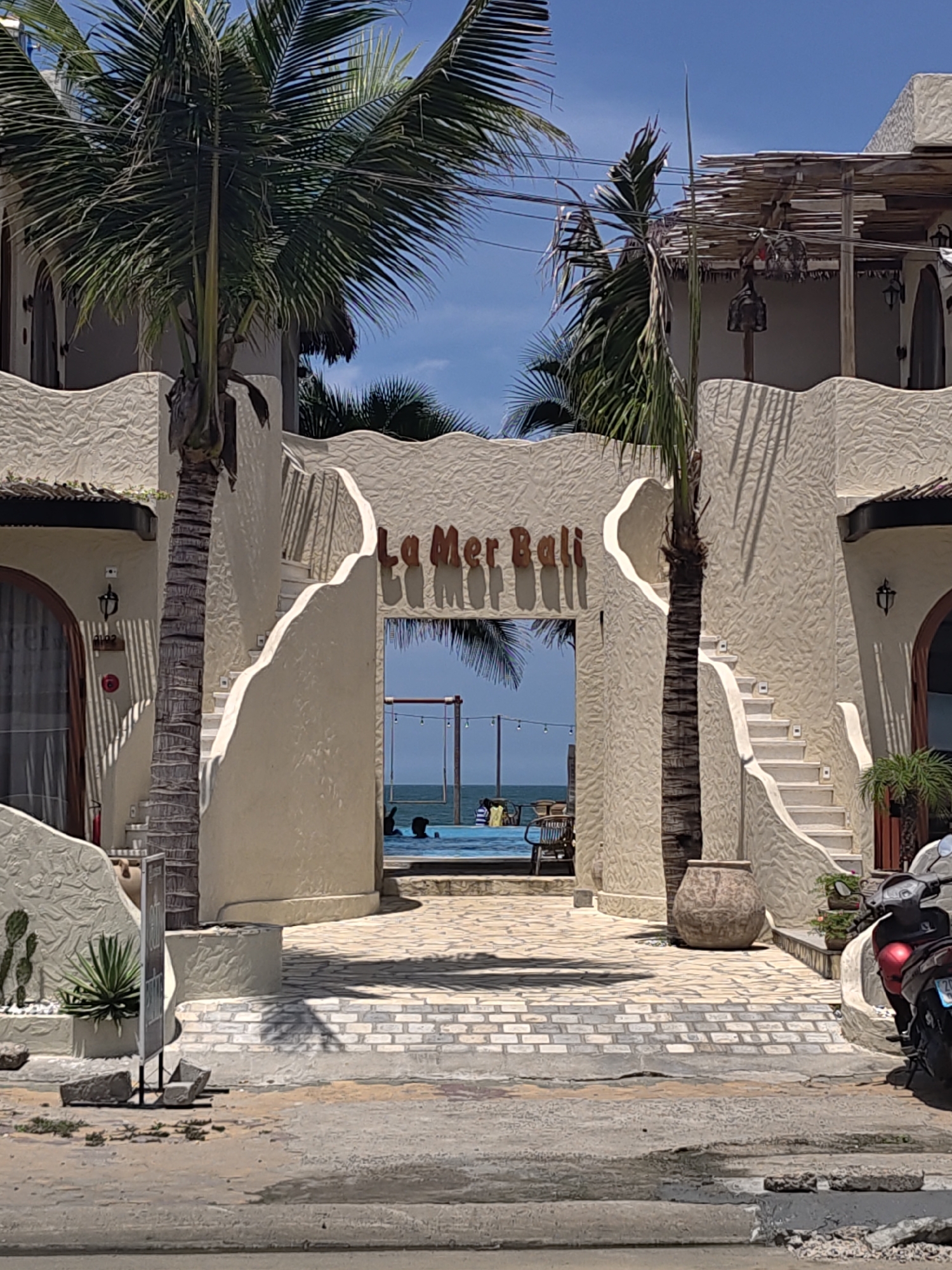 A daytime, eye-level, medium shot captures the entrance to a beachfront resort in Phu Thuy Ward, Vietnam, featuring a striking architectural design with creamy, textured walls and prominent palm trees. The central archway, adorned with the name "LA MER BALI" in stylized brown lettering, frames a view of a swimming pool and the ocean beyond. Two grand, curved staircases flank the archway, leading up to elevated sections of the building. A tall, slender palm tree stands directly in front of the archway, its fronds reaching towards a clear blue sky. To the left, a smaller palm tree partially obscures a building entrance with a sheer curtain. On the right, a red and black scooter is parked, partially visible, near a potted plant. In the background, a few people are visible in the swimming pool, and a rope swing hangs over the water. The ground is paved with a patterned arrangement of light-colored bricks. The overall impression is one of a serene, tropical getaway.