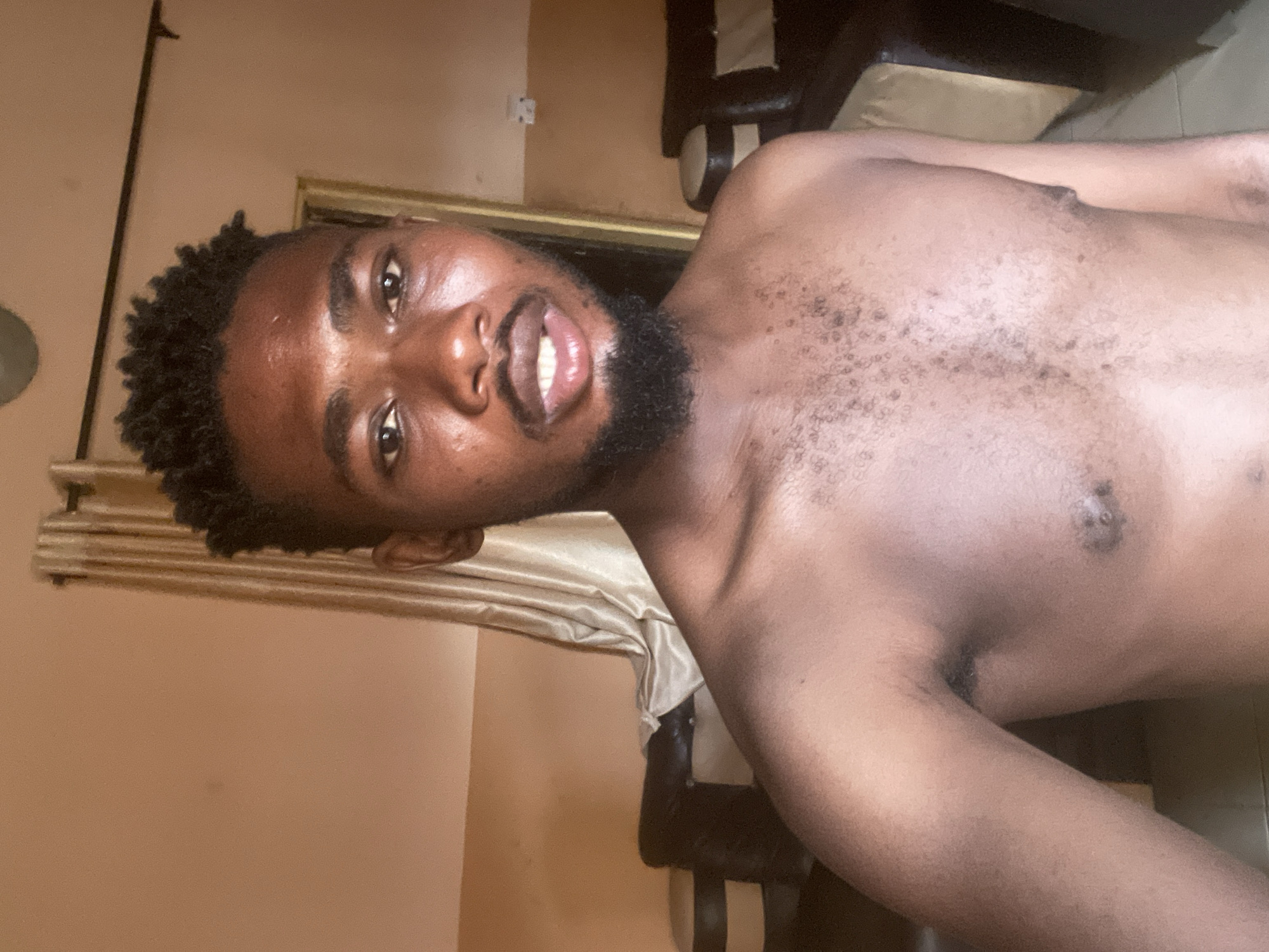 A shirtless man with a beard and short curly hair is posing for a selfie in a room with light brown walls. Curtains are partially visible behind him. A dark chair is also visible behind him. The location is suspected to be in Unguwar Gabas, Nigeria, but this cannot be definitively confirmed from the image alone.