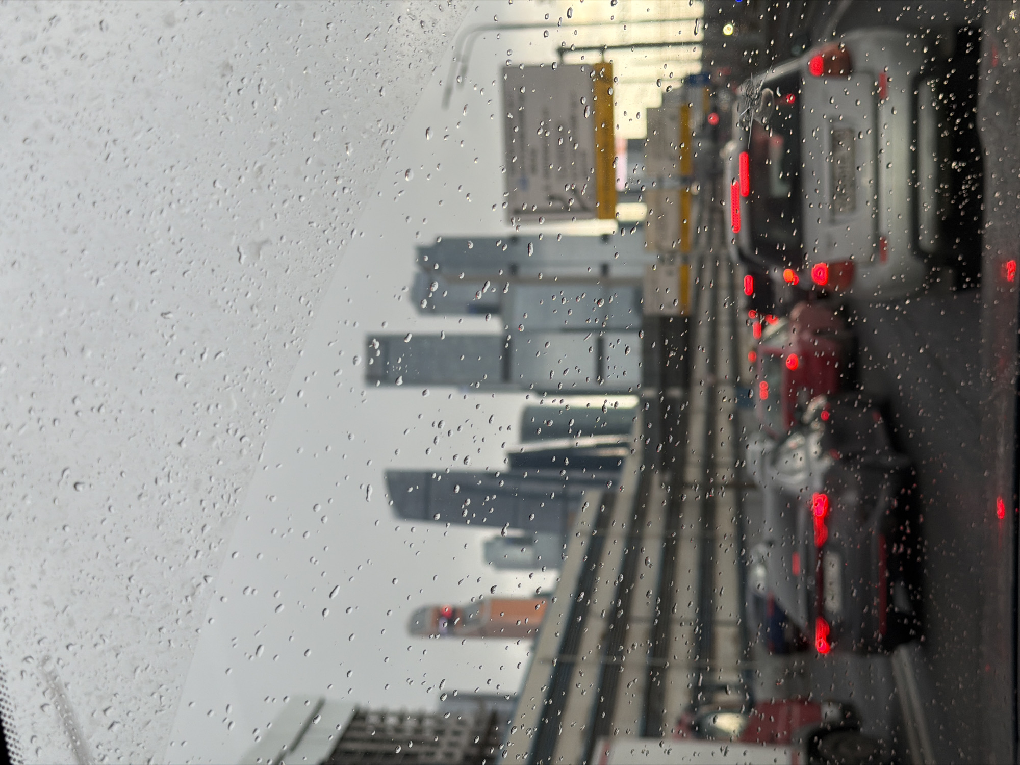 The image depicts a view through a rain-covered windshield, likely from inside a car. Outside, a cityscape is partially obscured by the rain and water droplets on the glass. The presence of high-rise buildings suggests an urban setting. A line of vehicles with illuminated taillights indicates traffic. The photograph was taken in Khimki, Russia.