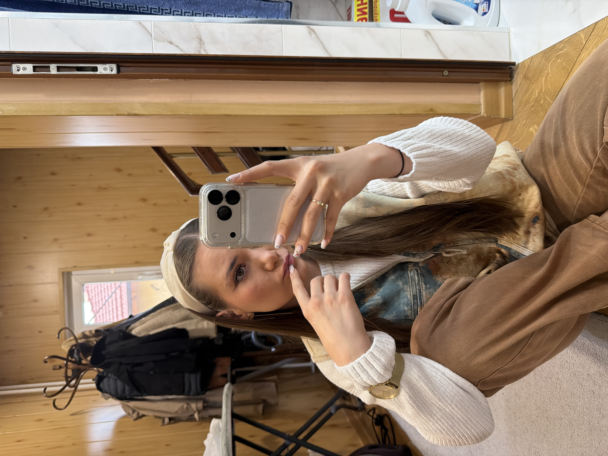 A young woman is captured taking a mirror selfie indoors in Buhovo, Bulgaria. She is holding a silver iPhone with a clear case in her right hand, while her left hand is touching her lips. She has long brown hair, a white headband, and is wearing a white ribbed sweater, brown pants, and a distressed denim jacket. A gold watch adorns her left wrist, and a gold ring is on her right hand. Her nails feature white tips with small black polka dots.
The setting appears to be a room with warm wooden paneling on the walls. In the upper background, there's a white tiled surface, possibly a counter or backsplash, where a white bottle with partially visible Cyrillic text ("БИОЛОГИЧ...") is placed. Above this counter, a metal lock mechanism is visible on a wooden frame. The mirror's reflection reveals a window looking out onto a building with a red-tiled roof, indicating daytime. Below the window, a wooden coat rack holds several dark coats. The overall impression is that of a cozy, possibly rustic, home interior.