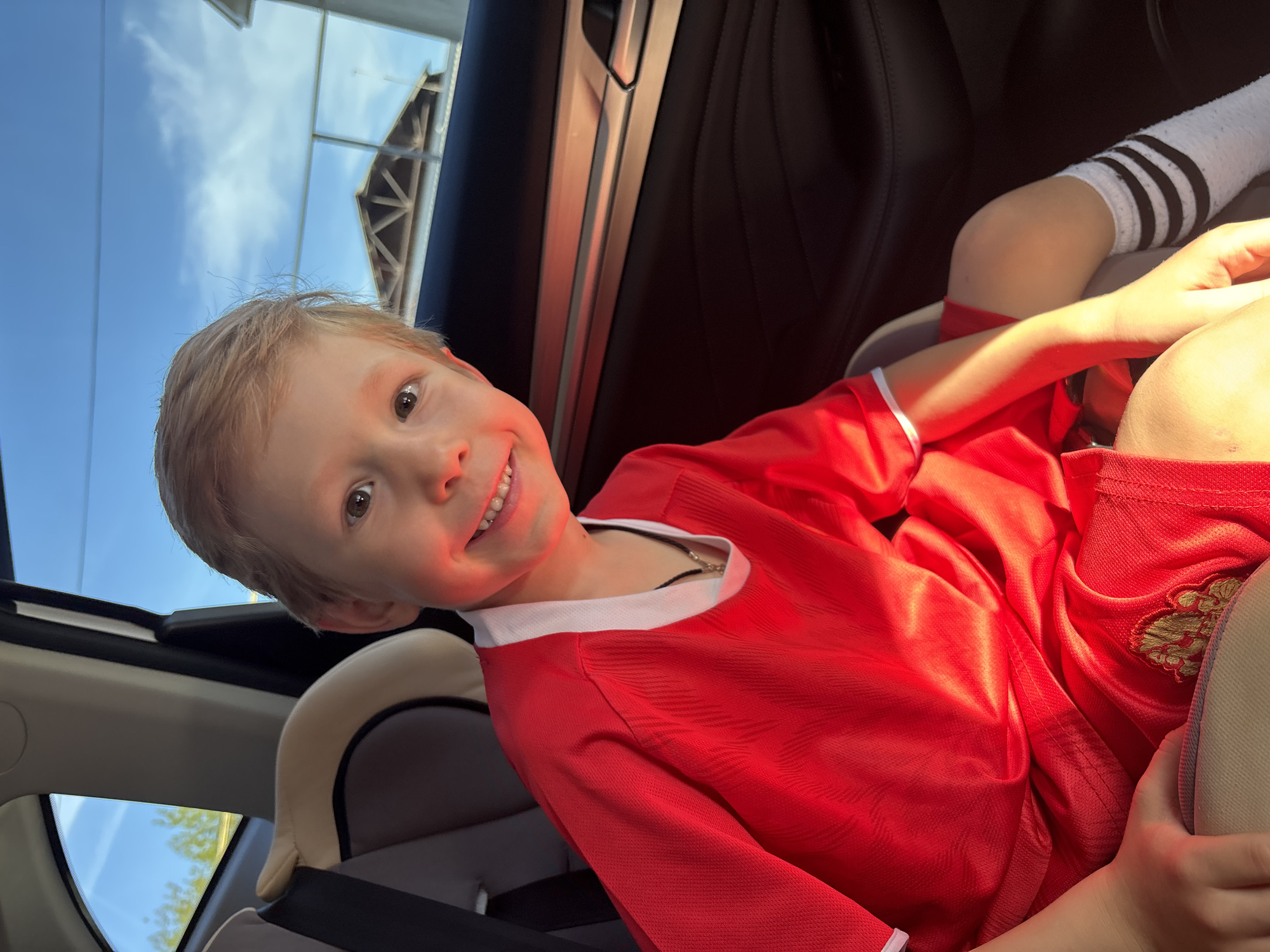 A young child with light blonde hair is smiling in a car. The child is wearing a red jersey with white trim around the collar and sleeves and is buckled into a car seat. Part of another child's arm, clad in a white sock with black stripes, is visible. The car has a sunroof with a view of the sky. The exact location in the city of Chaikovsky, Russia, cannot be determined from the image.