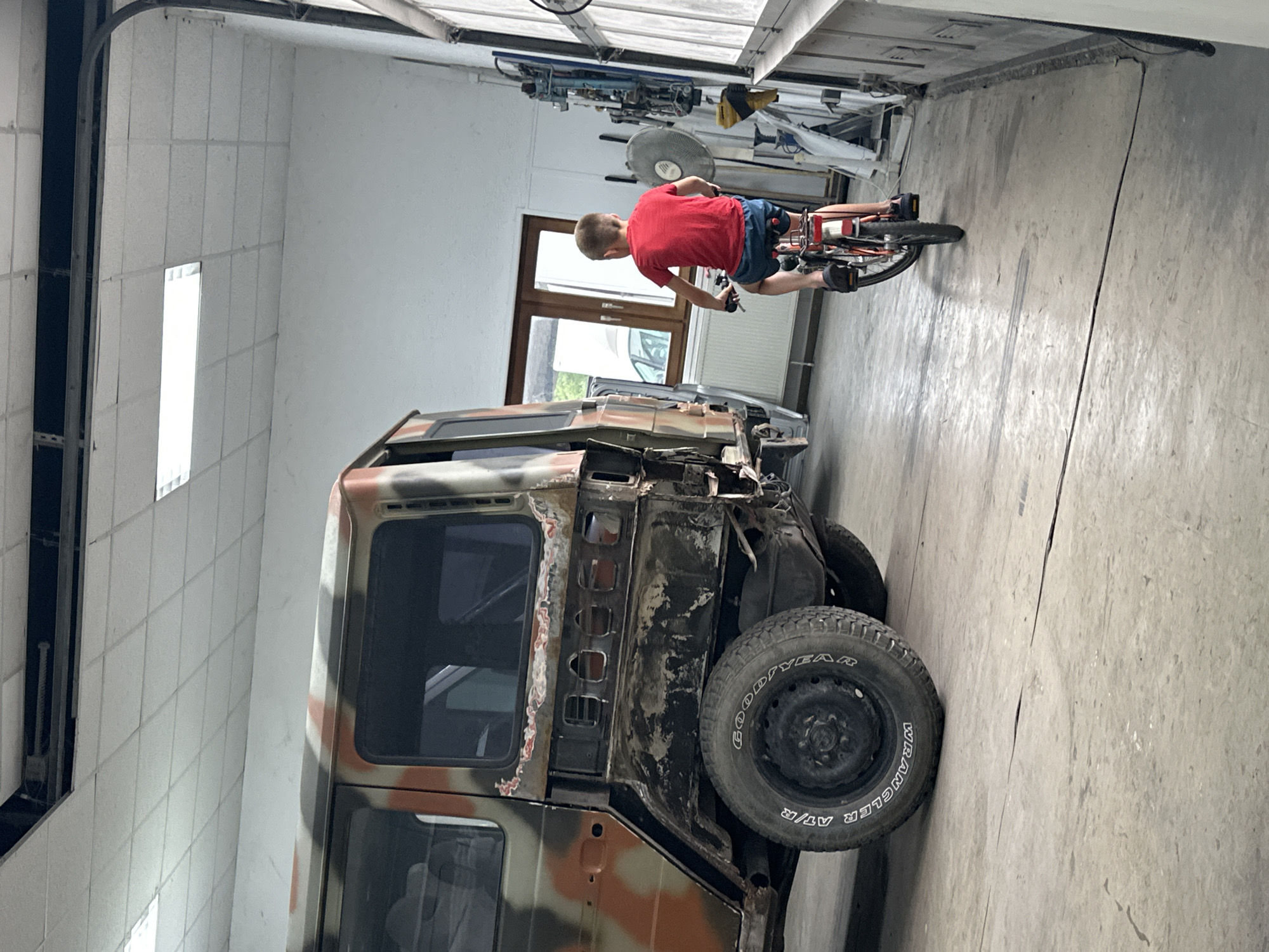 In an indoor garage, possibly a workshop, in the city of Mainz, Germany, a young boy is riding a bicycle. He is wearing a red t-shirt and blue shorts. The bicycle is red and has a white rear fender. The boy is facing away from the camera. To his left is the rear portion of a large, weathered, camouflage-painted vehicle, possibly a military truck or SUV, which appears to be under repair or restoration. The vehicle has large tires and its bodywork shows signs of rust and damage. The garage floor is concrete. In the background, there are windows, industrial lighting fixtures on the ceiling, and various tools and equipment mounted on the wall.