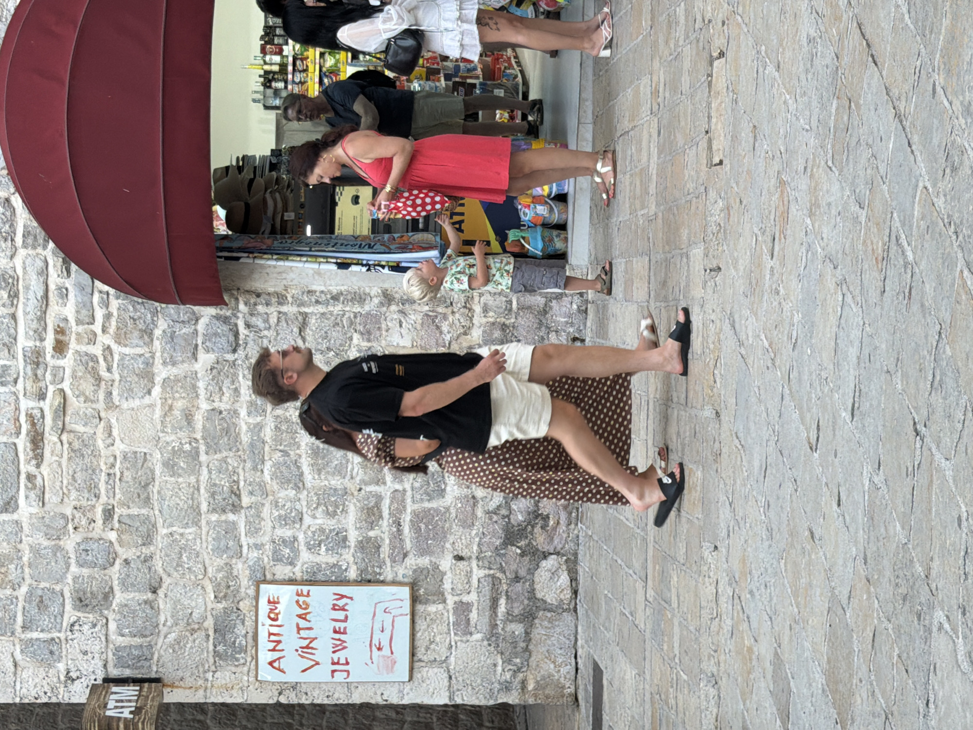 The image depicts a street scene in Budva, Montenegro, outside a shop with a red awning. A man in a black t-shirt and light-colored shorts walks past a sign that reads "ANTIQUE VINTAGE JEWELRY." He is barefoot with black slides on his feet. To his left, a woman in a brown polka dot dress walks in the same direction. A young boy in a patterned shirt and grey shorts stands by the entrance of the shop, interacting with a woman in a bright pink dress. The woman in pink is holding a red bag with white polka dots. Several other people are visible in the background, including a man in a dark t-shirt and shorts, and a woman in a white dress. The ground is paved with cobblestones. The shop appears to sell hats and other merchandise. An ATM sign is visible on the stone wall to the left of the image.