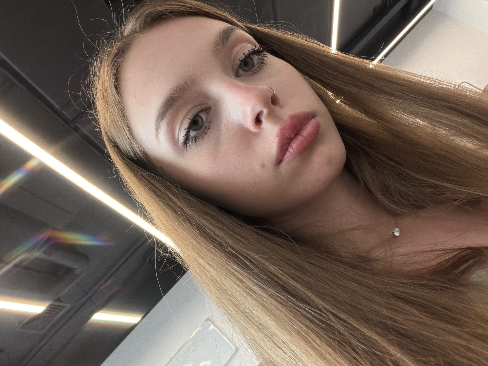 A close-up, high-angle, indoor shot captures a young woman with long, light brown hair in a selfie. Her face is angled downwards and slightly to the left. She has grey eyes, dark eyelashes, and light pink lipstick. A small, silver nose ring is visible on her right nostril. She is wearing a delicate silver necklace with a single clear stone. The background features dark, textured ceiling panels with bright, linear light fixtures. A faint rainbow-like refraction is visible on the ceiling to the left. A white board with faint writing is partially visible in the upper left corner. The lighting is bright and directed, creating highlights on her hair and skin.