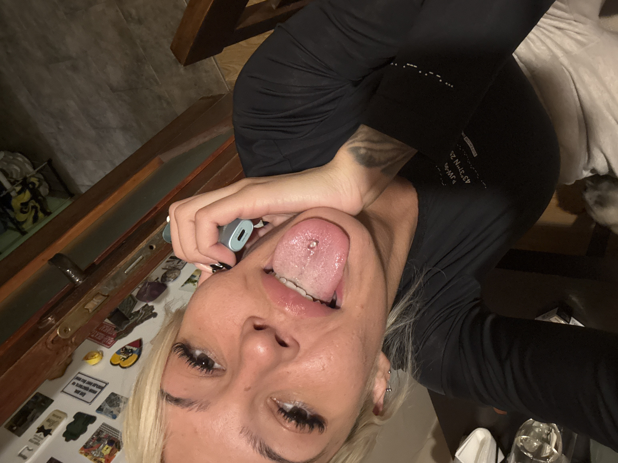 A close-up, tilted shot features a light-skinned individual with blonde hair, dark eyelashes, and a pierced tongue. The person is holding a small, light-blue object near their mouth. The background includes a cluttered surface with various magnets and stickers. To the right of the person, there is a glimpse of a black garment with text, possibly coordinates, and a partial view of a white object. At the bottom right, there is a clear glass container.