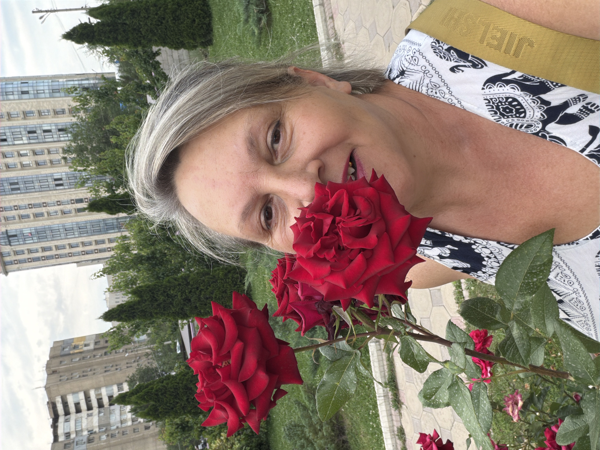 A middle-aged woman with graying blonde hair takes a selfie with a bouquet of red roses in the foreground. She is smiling, and a few of the rose petals are obscuring her mouth. She wears a white top with a black elephant pattern and a yellow strap across her shoulder. In the background, there are tall, modern buildings and manicured green bushes, suggesting an urban park setting. The sky is overcast.