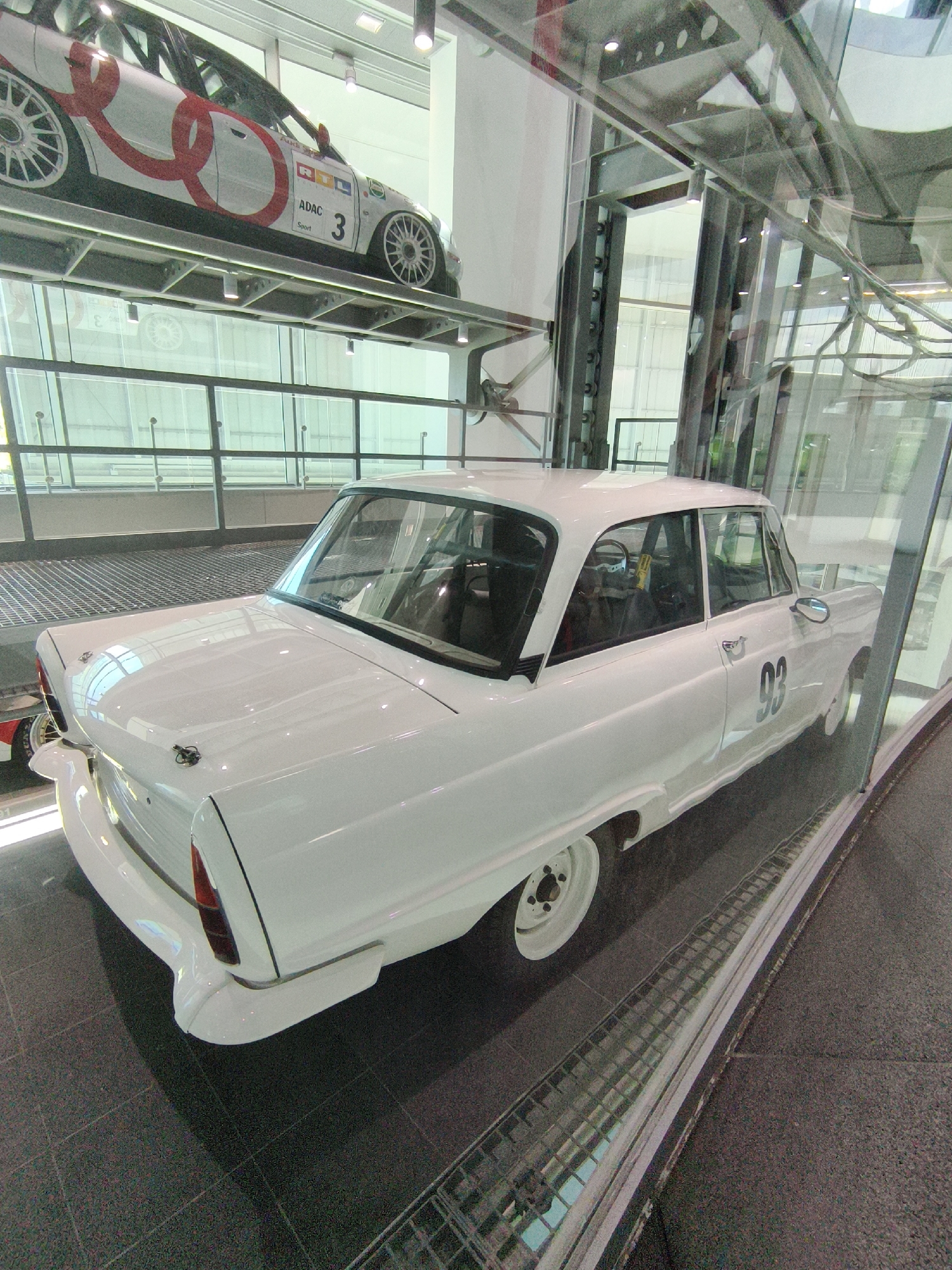 The image shows a white vintage Audi car displayed in a modern exhibition hall, likely the Audi Forum Ingolstadt in Ingolstadt, Germany. The car is positioned behind a glass enclosure. Above, another Audi race car is visible on a raised platform. The exhibition hall features industrial-style architecture with metal beams and glass walls. The lighting is bright and even, typical of a museum or showroom. The white car has racing numbers, "93," on its side. The foreground shows the dark tiled floor and a metal grating.