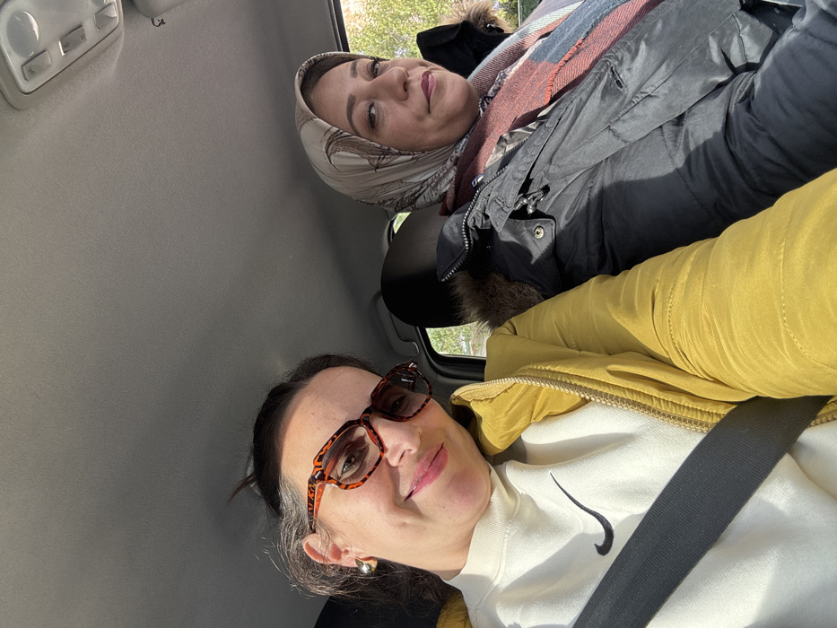 The image captures two women inside what appears to be a car, photographed from a low angle looking upwards, and rotated clockwise.
In the upper right portion of the frame, the first woman is visible, sitting in the passenger seat further back. She is wearing a light-colored headscarf (hijab) with a subtle pattern, possibly in shades of beige or off-white. Her eyes are light in color, and she has a gentle smile on her face. She is dressed in a dark, possibly black, winter jacket with a fluffy, fur-lined collar or hood visible around her neck and shoulders. A plaid scarf, featuring warm tones like red, orange, and possibly some blue or green, is wrapped around her neck, tucked into her jacket.
In the lower left foreground, closer to the camera, is the second woman. She is wearing brown, tortoiseshell-patterned eyeglasses. Her dark hair is pulled back, and she has a soft, closed-mouth smile. She is wearing a white sweatshirt with a prominent black Nike swoosh logo on the front. Over the sweatshirt, she wears a warm, mustard yellow or olive green puffer jacket. A dark seatbelt crosses diagonally over her chest and shoulder. A small, round gold earring is visible on her left ear.
The setting is clearly the interior of a car, indicated by the light grey headliner (ceiling) visible across the top and left side of the image. An overhead console with control buttons can be seen near the top left corner. Through the car windows, some blurred greenery and bright daylight are visible, suggesting it is daytime and possibly a clear day. Both women are looking towards the camera, indicating either a selfie or a photograph taken by a third person in the vehicle. Their expressions suggest a calm and pleasant mood.