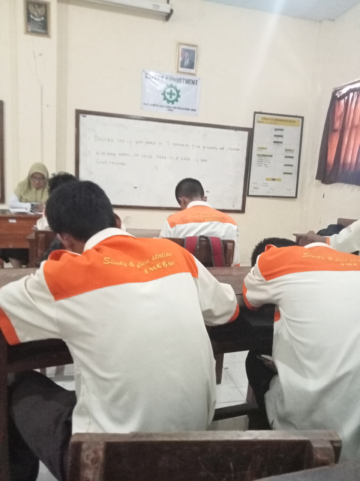 The image depicts an indoor scene, likely a classroom in Secang, Indonesia, where students are seated at desks. The focus is on the backs of several students wearing white shirts with orange accents. The shirts have embroidered text that reads "Study & fun station SMK Bisa".
On the wall behind the students, there is a whiteboard with handwritten text and a diagram. The text on the whiteboard appears to be an assignment. To the right of the whiteboard, a framed chart labeled "STRUKTUR ORGANISASI KELAS" (Class Organizational Structure) is displayed.
In the background, a teacher wearing a light-colored hijab is visible at a desk, seemingly observing the students or engaged in her own work. A framed photograph of a man in a suit hangs on the wall to the left of the whiteboard, and a banner with a green cross symbol and text that includes "COMMITMENT" is also present. The room appears to be lit by natural light entering from the right side, where curtains are visible. The overall atmosphere suggests a typical school day.