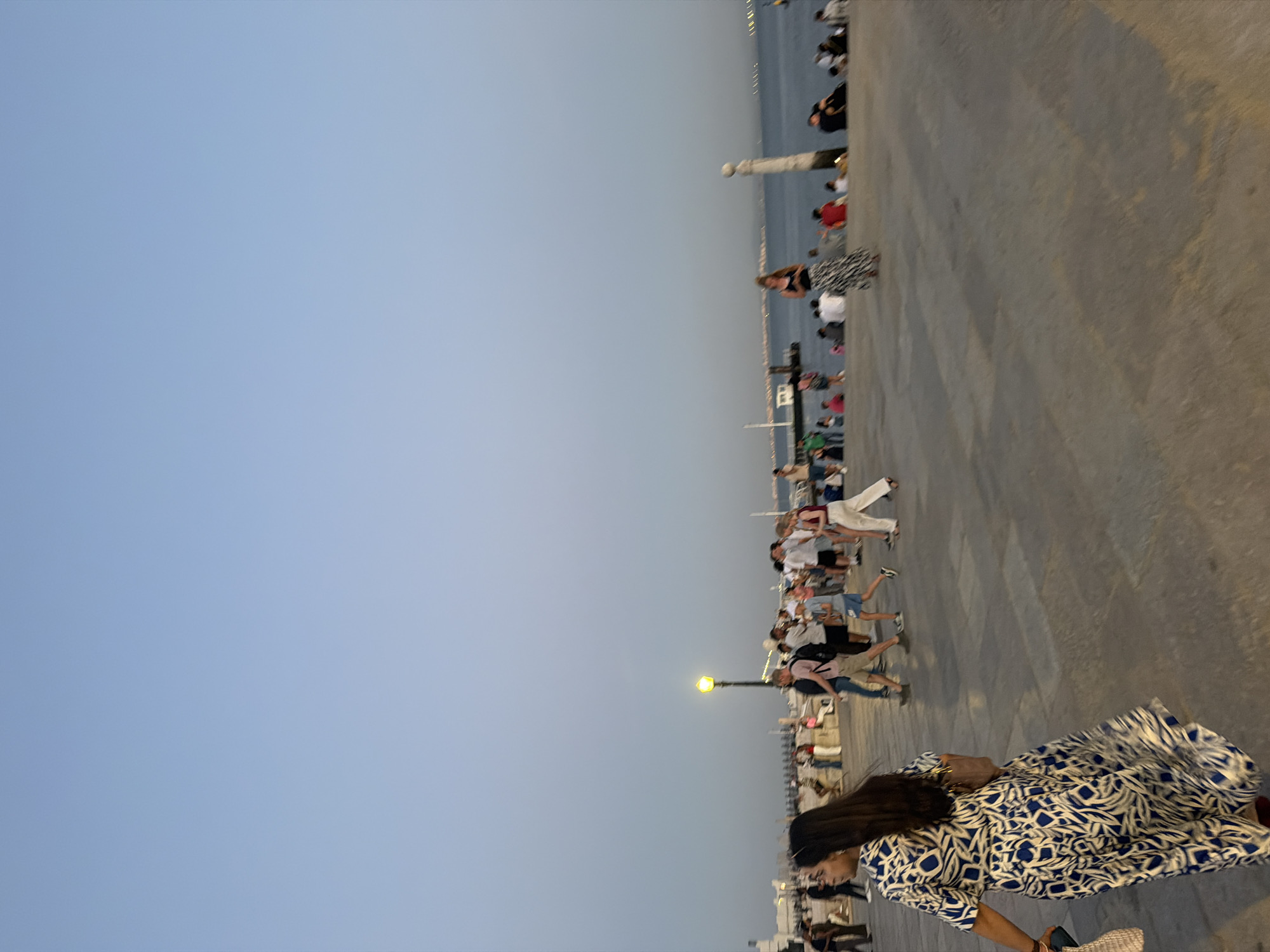 A street-level, medium shot captures a promenade in Lisbon, Portugal, with the Tagus River visible in the background. The sky is a clear, pale blue, indicative of early evening or late afternoon light. A paved promenade, made of light gray stone, occupies the foreground and midground. Several people are walking and standing on the promenade. On the left, a woman with long dark hair, wearing a blue and white patterned dress, walks away from the camera, carrying a tote bag. To her right, a group of people are gathered and walking, many appearing to be tourists. They are dressed in casual summer clothing. A prominent yellow lamppost is positioned in the midground, illuminated. Further back, the Tagus River stretches across the horizon, with a few boats and distant city lights visible. A cylindrical stone monument stands near the water on the right side of the frame. The overall atmosphere suggests a popular public space in Lisbon during pleasant weather.