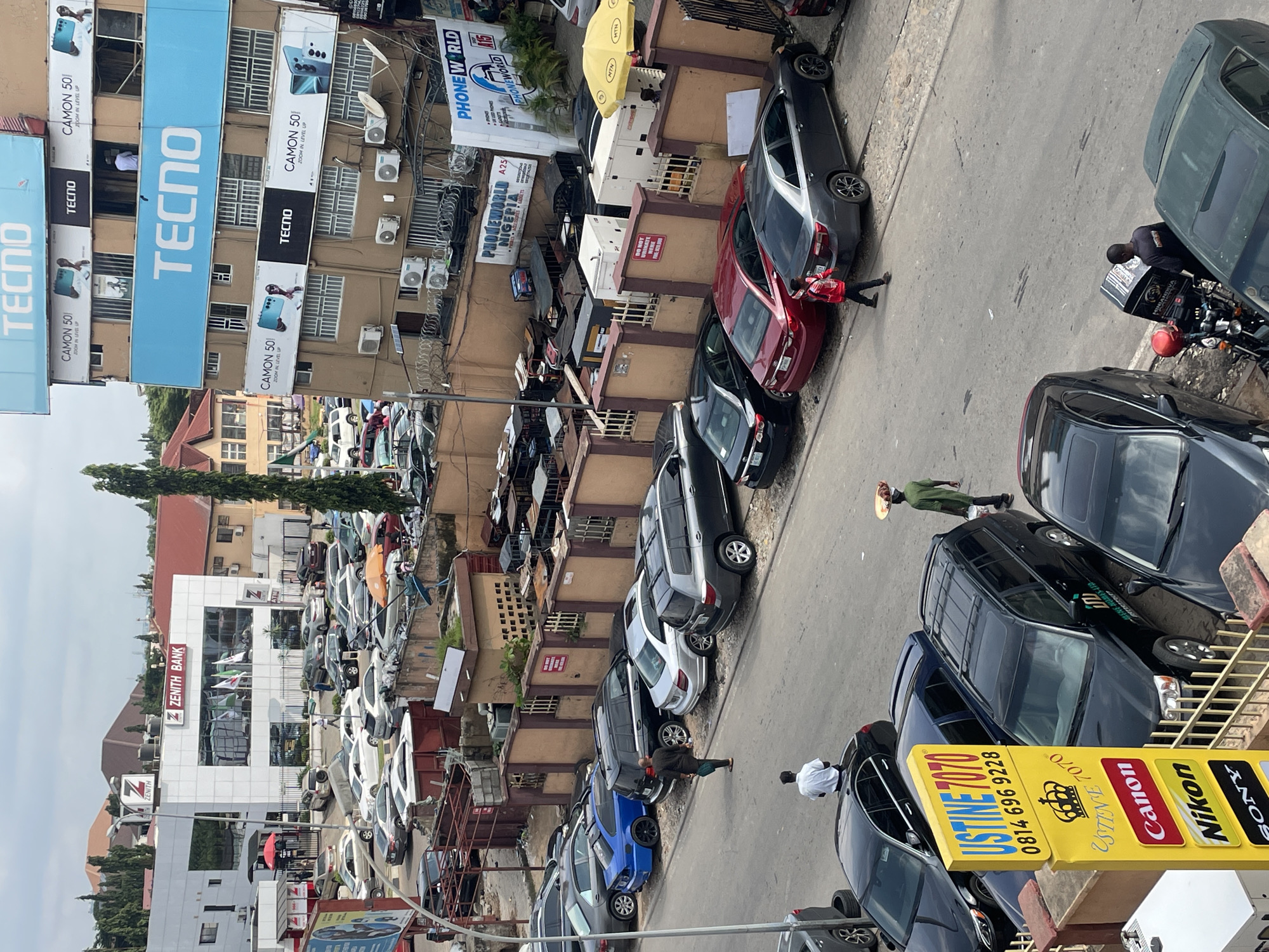 The image depicts a street scene in Abuja, Nigeria, likely in a commercial district given the presence of businesses and vehicles. Several buildings are visible, with prominent signage for "ZENITH BANK" and "TECNO," suggesting a focus on banking and telecommunications/electronics. A multi-story building branded with "TECNO" is adorned with large advertisements for "CAMON 50." Other businesses like "PHONEWORLD NIGERIA" are also present.
The street is lined with parked cars, including sedans, SUVs, and at least one blue sports car. Some vehicles appear to be in a parking lot, while others are parked along the roadside. The weather seems overcast.
Several individuals are present in the scene. One person is seen walking away from the camera, carrying a tray of items on their head, a common sight for street vendors in Nigeria. Another individual in a green shirt is also walking. A person in a white shirt is seated on a car. A person in a red jacket is walking on the street. A person is standing near a motorcycle with a delivery box. A man in a dark shirt and pants is walking on the street, carrying a bag. People are also seen near the entrance of Zenith Bank.
The environment includes pavements, road surfaces, streetlights, and various architectural elements of the buildings. The overall impression is of a bustling urban environment with commercial activity. A yellow sign with "USTINE 7070" and logos for Canon, Nikon, and Sony is prominently displayed, likely advertising a service or product related to electronics or photography.