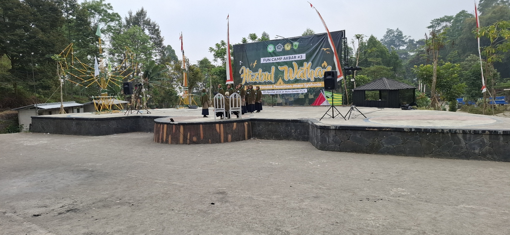 The image depicts an outdoor stage setup in a park, likely in the city of Karang, Indonesia. The stage is elevated and has a stone-like textured base. Behind the stage, a large green banner with text in a script font reads "Hizbul Wathan" and also displays "FUN CAMP AKBAR #3" and "Pondok Pesantren Imam Syuhodo, 20 Agustus 2026 at Watu Gambir Park". Several individuals, appearing to be young women or girls dressed in brown uniforms and headscarves, are standing on the stage, seemingly preparing for or participating in an event. To the left of the stage, a large, intricate bamboo structure, possibly a decoration or part of a performance, is visible, along with some buildings in the background. Two loudspeakers are positioned on stands on the stage. The environment is lush with trees, indicating a natural or park-like setting. Flags are present, including a red and white Indonesian flag. A small, dark-colored gazebo-like structure is situated to the right of the stage. The ground in front of the stage is paved with concrete.