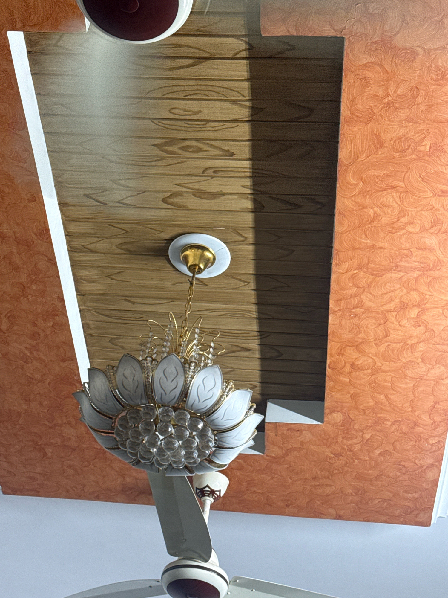 The image shows a close-up of a ceiling with a decorative chandelier and a ceiling fan. The ceiling features a combination of wood-textured panels and textured orange panels. A shadow is cast across the wood-textured panels. The chandelier is ornate, with white petal-like accents and clear crystal-like beads. The ceiling fan has white blades and a decorative base. The image was taken indoors, likely in a residential setting. The location is identified as Gujrat, Pakistan.