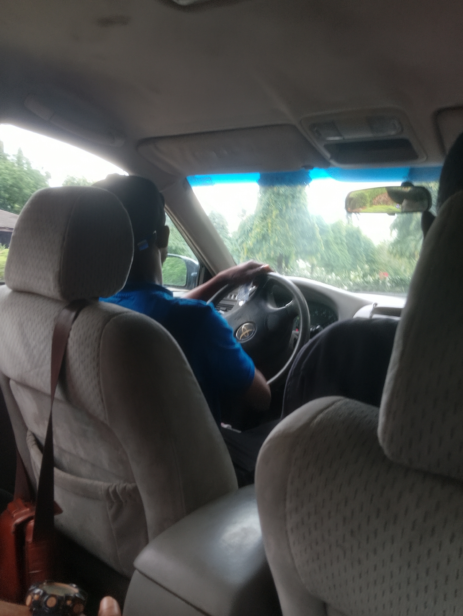 An interior shot from the back seat of a car shows a male driver in a blue shirt. He is wearing headphones and holding the steering wheel, which has a Toyota logo. Outside the car, lush green trees are visible through the windshield and side windows. The interior of the car features beige-colored seats. This scene appears to be in or around Iju, Nigeria, based on the vegetation and general appearance.
