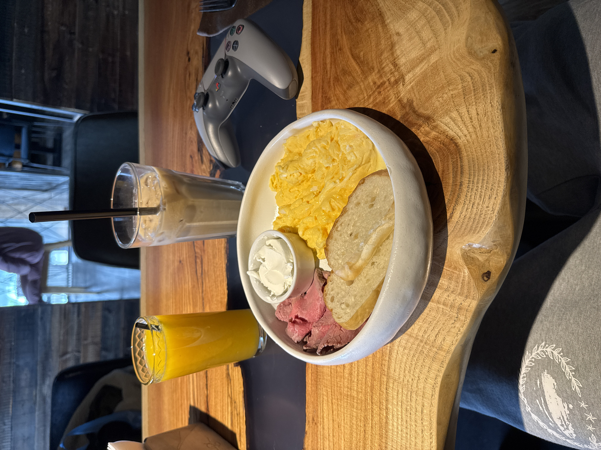 A table scene, likely a cafe or restaurant, shows a wooden table top with a plate of food. The plate contains scrambled eggs, sliced bread, thinly sliced meat, and a small dish of white cream or cheese. Next to the plate, there's a glass of orange juice and a tall glass with a beverage that appears to be iced coffee. A gray PlayStation controller is also on the table. The location of the image is Khimki, Russia.