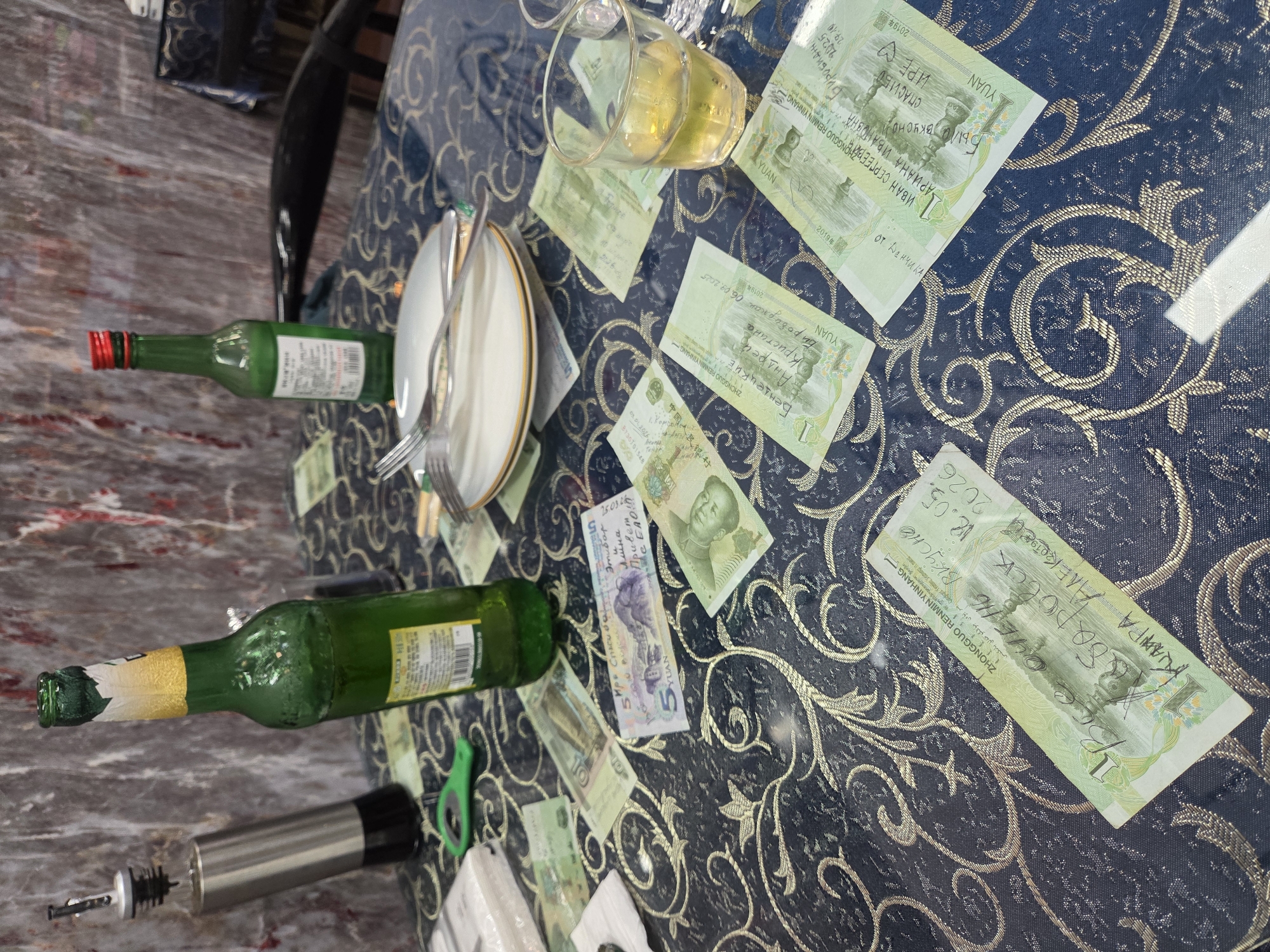 The image, rotated 90 degrees clockwise, presents a tabletop scene, likely following a meal or social gathering. A dark blue tablecloth adorned with ornate gold patterns covers the table.
Scattered across the tablecloth are multiple Chinese Yuan banknotes of 1, 5, 10, and 50 Yuan denominations. A significant detail is the presence of handwritten messages in Cyrillic script (Russian) on many of these notes. Visible text includes "YUAN" and "ZHONGGUO RENMIN YINHANG" (People's Bank of China). The handwritten inscriptions convey expressions such as "Спасибo" (Thank you), "ОЧЕНЬ Вкусно" (VERY delicious), and "БЫЛО ВКУСНО СПАСИБО ИРЕ" (IT WAS DELICIOUS THANK YOU IRA). Names like "Айна," "Этибар," "Иван Сергеев," "Ариана Ивановна," and "от Денчика" (from Denchik) are also visible. Some notes bear specific dates and cities, including "Биробиджан 06.09.2025" and "Хабаровск 18.05.2026," as well as "КРАМПА АЛЕКОС 2026." Dates like "2019年" (Year 2019) and a serial number "8730701548" can be seen on some notes.
Other objects on the table include two green glass bottles, one with a red cap and another with a gold and white label, suggesting beverages. A glass containing a light yellow liquid, possibly an alcoholic drink, is present. A stack of white plates with gold rims and silverware (forks) are arranged nearby. A metallic and clear glass wine stopper/pourer and small green plastic clips are also on the table. The background features a light-colored, possibly marbled, surface.
The scene implies a casual dining event, possibly in Tongjiang, China, where guests, potentially from Russian-speaking areas, have left personalized notes on the local currency as a memento or sign of appreciation.