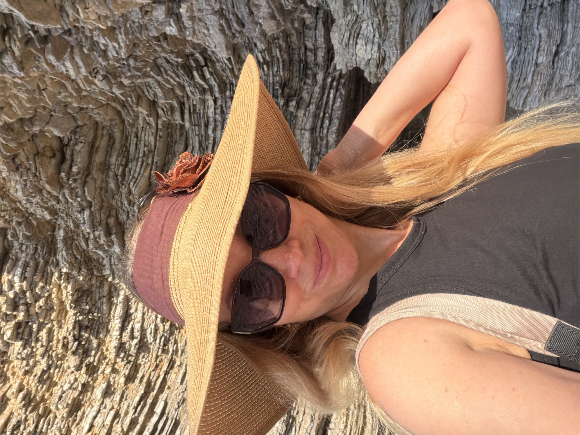 A middle-aged woman wearing a wide-brimmed sun hat, sunglasses, and a dark-colored top is reclining outdoors. She is positioned against a background of sedimentary rock formations. The woman appears to be posing for the photo. This scene is likely situated in the coastal area of Budva, Montenegro, where similar rock formations are prevalent.