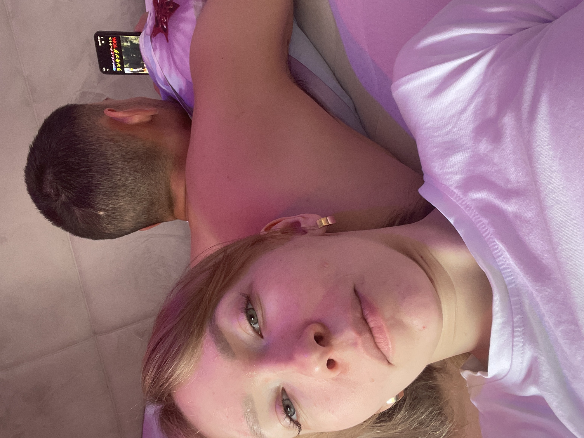 A selfie captures a woman with fair skin, light brown hair, and light-colored eyes. She is wearing hoop earrings and a white shirt. Behind her, a man with short dark hair is lying down, looking at a smartphone. The screen displays an image with text in a non-Latin script. They are both lying on a surface that appears to be a bed or couch covered with fabric. The lighting in the image has a pink hue. There is no information to specify the location in Perm, Russia.