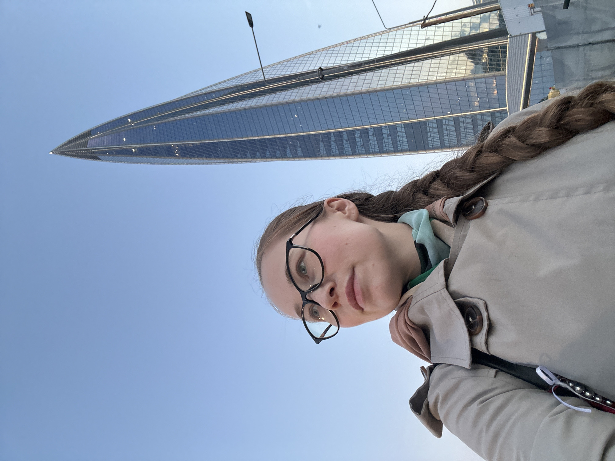A young woman with brown hair braided down her back, wearing glasses and a tan trench coat, poses in front of the Lakhta Center skyscraper in Saint Petersburg, Russia. The building is constructed of glass and steel, with a needle-like spire. A street lamp and parts of adjacent structures are visible in the upper portion of the frame. The background is a clear blue sky.