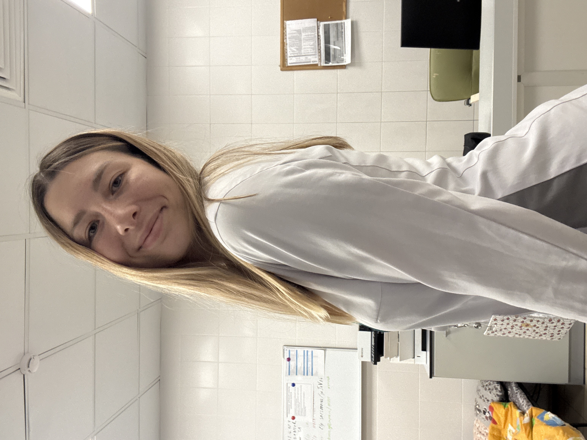 A young woman with long blonde hair, wearing a light-colored uniform top, poses for a selfie in what appears to be a medical or laboratory setting. She has a slight smile and is looking towards the camera. The background features tiled walls, a suspended ceiling with lighting panels, and a whiteboard with some writing. There are also cabinets, a desk with a laptop, and a bulletin board visible. The overall impression is that of a professional or student in a healthcare environment. The image does not contain any information or visual cues that would allow for its location to be identified as Moscow, Russia.